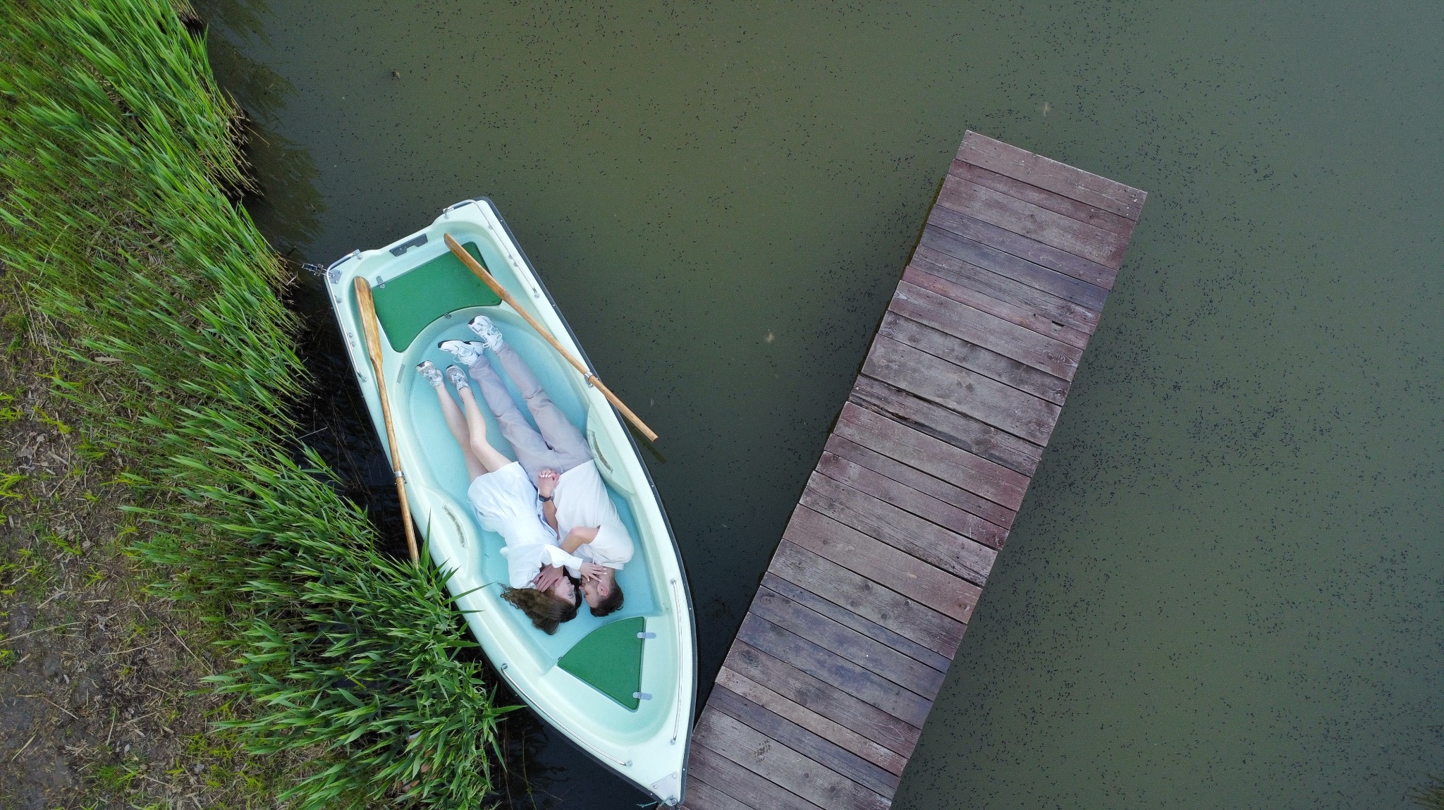 Para leżąca w łódce przy drewnianym pomoście, widok z góry na zieloną wodę i trzciny.