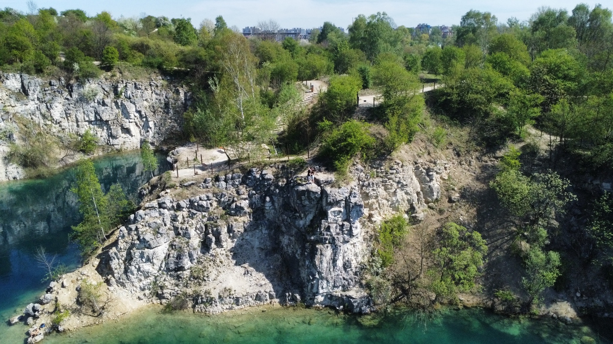Para na skalnym klifie nad turkusowym jeziorem w dawnym kamieniołomie; w tle zieleń drzew i zabudowania miejskie.