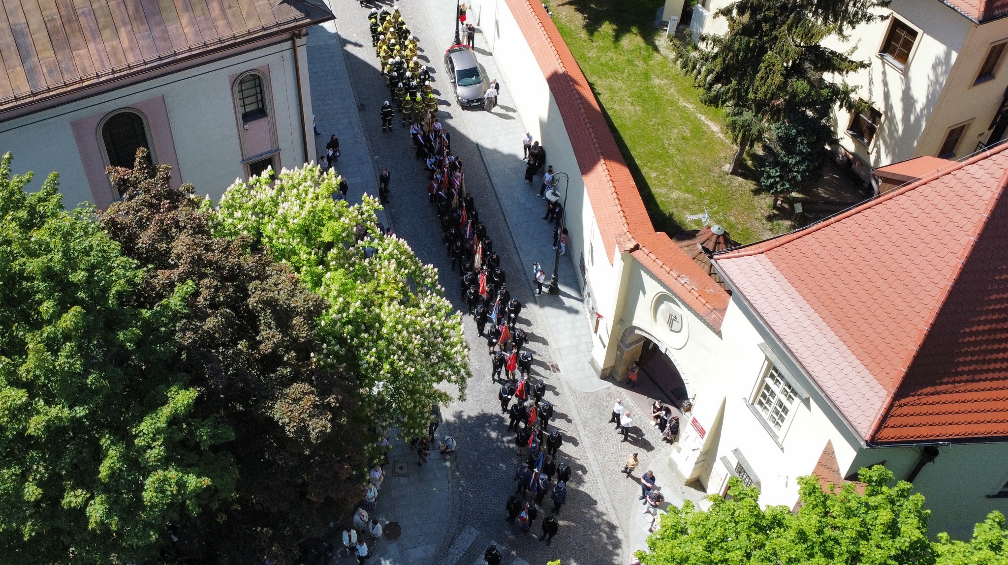 Widok z góry na ulicę z budynkami i drzewami, gdzie maszeruje długa kolumna strażaków i osób z flagami, a na chodnikach stoją obserwatorzy.