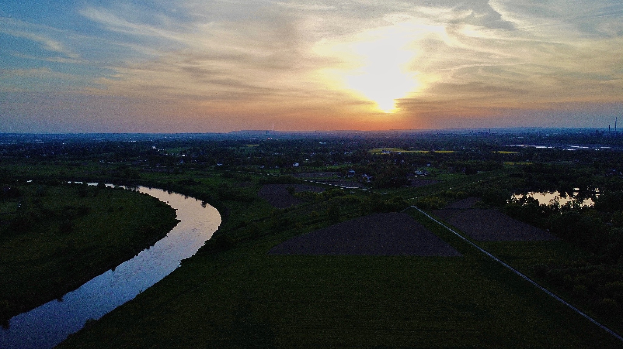 Panoramiczny widok z drona na zakole rzeki, pola uprawne i odległe zabudowania miejskie o zachodzie słońca.