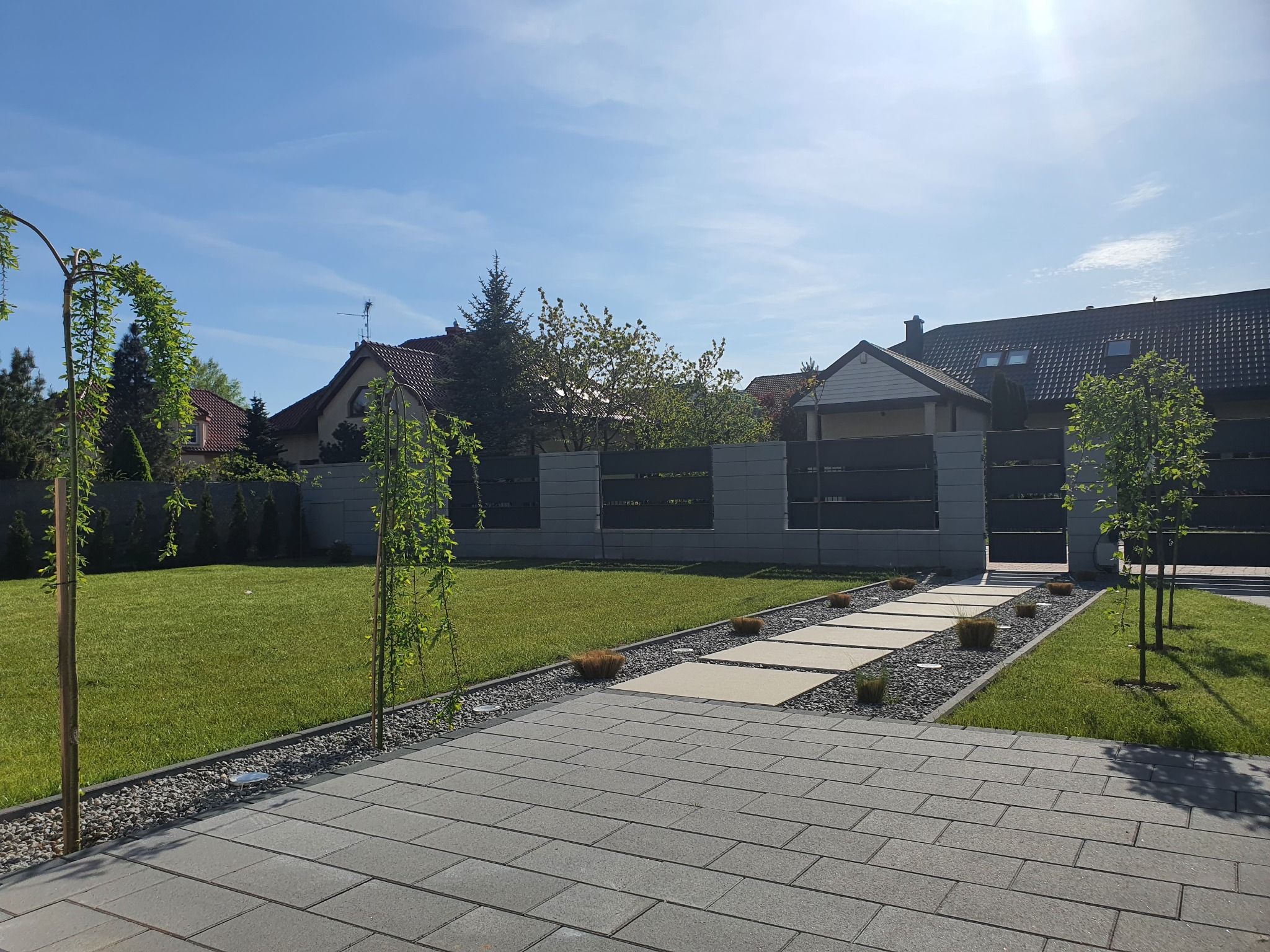 Nowoczesny ogród z równo przystrzyżonym trawnikiem, ścieżką z płyt i ozdobnymi drzewkami. Widoczny fragment domu i ogrodzenie w tle, słoneczny dzień.