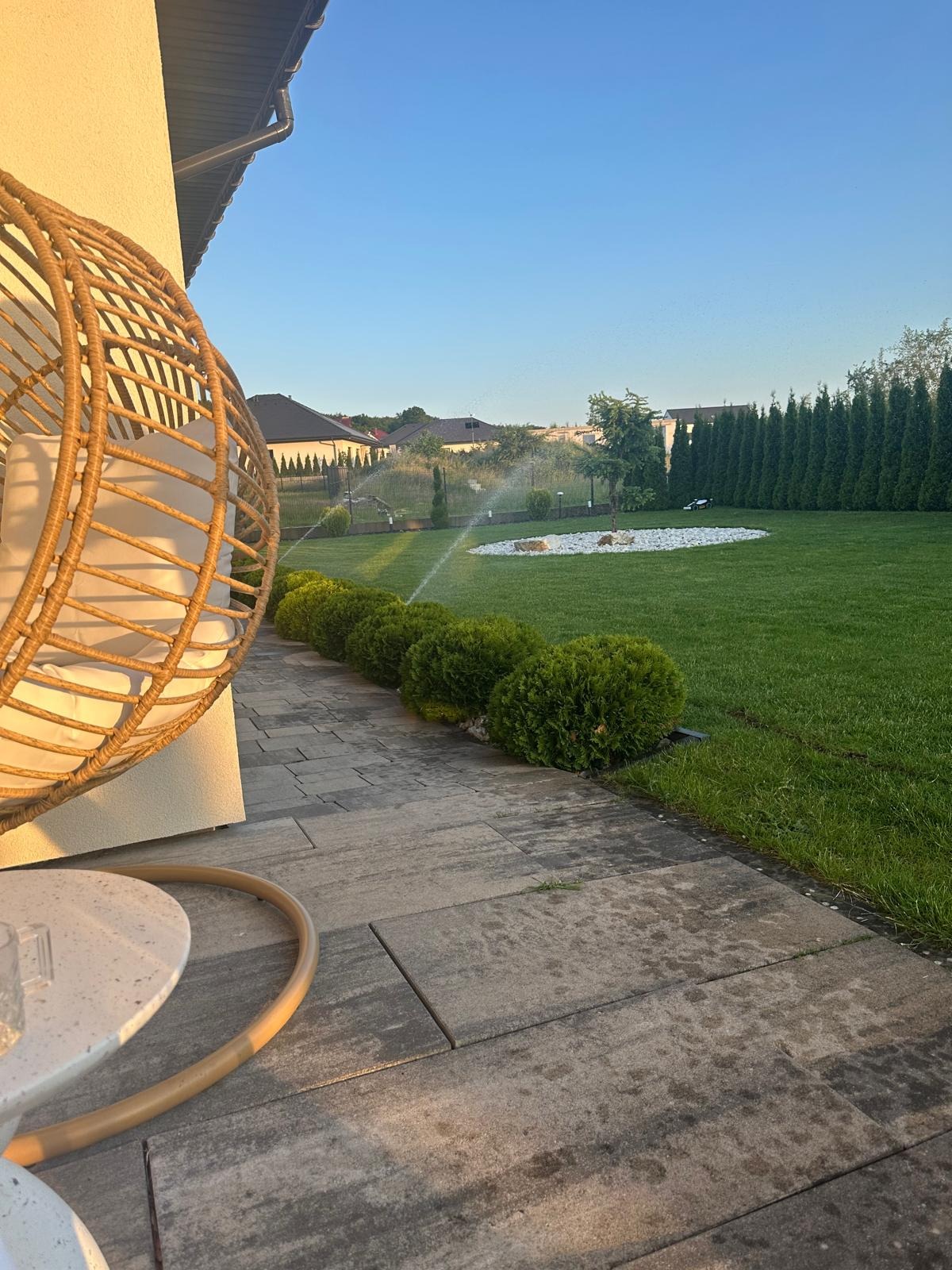 Automatyczne nawadnianie trawnika z dyszami, widok z tarasu z rattanowym fotelem. Zadbany ogród z równo przystrzyżonymi krzewami i trawą.