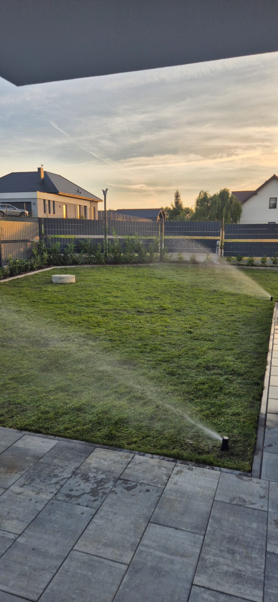 Automatyczny system nawadniania trawnika w ogrodzie, zraszacz w akcji, w tle dom jednorodzinny i ogrodzenie panelowe. Zadbany trawnik, wieczorne słońce.