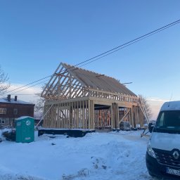 Dachy Dela - Wznoszenie szkieletu drewnianego domu w zimowej scenerii, widoczny przenośny WC i samochód dostawczy na pierwszym planie.