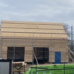 Dachy Dela - Konstrukcja domu w trakcie budowy, widoczne drewniane bale i rusztowania, z boku leżą materiały budowlane.