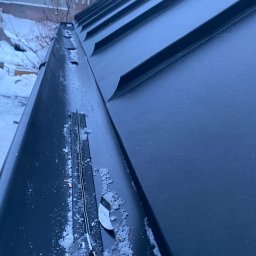 Dachy Dela - Nowy, ciemny dach z blachy trapezowej pokryty lekkim śniegiem, widok z góry na krawędź dachu i rynnę z widocznymi elementami mocującymi.