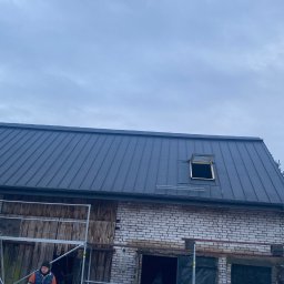 Dachy Dela - Budynek z nowym, ciemnym dachem z blachy na rąbek stojący, otwartym oknem dachowym, fragmentem rusztowania i pracownikiem w dole, w tle pochmurne niebo.