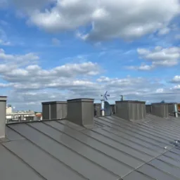 Szary metalowy dach z kominami i anteną satelitarną na tle błękitnego nieba z obłokami.