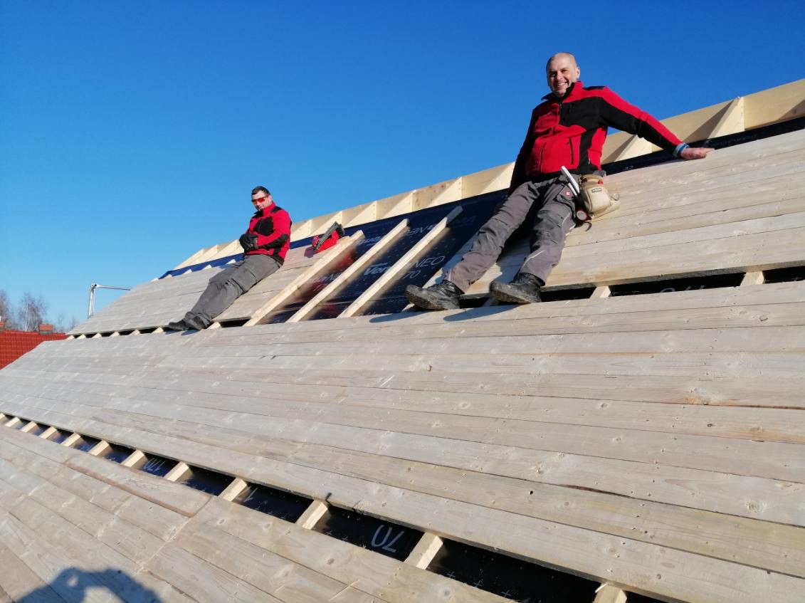 Dwóch dekarzy w czerwonych kurtkach siedzi na drewnianej konstrukcji dachu pokrytej czarną membraną, w tle błękitne niebo.