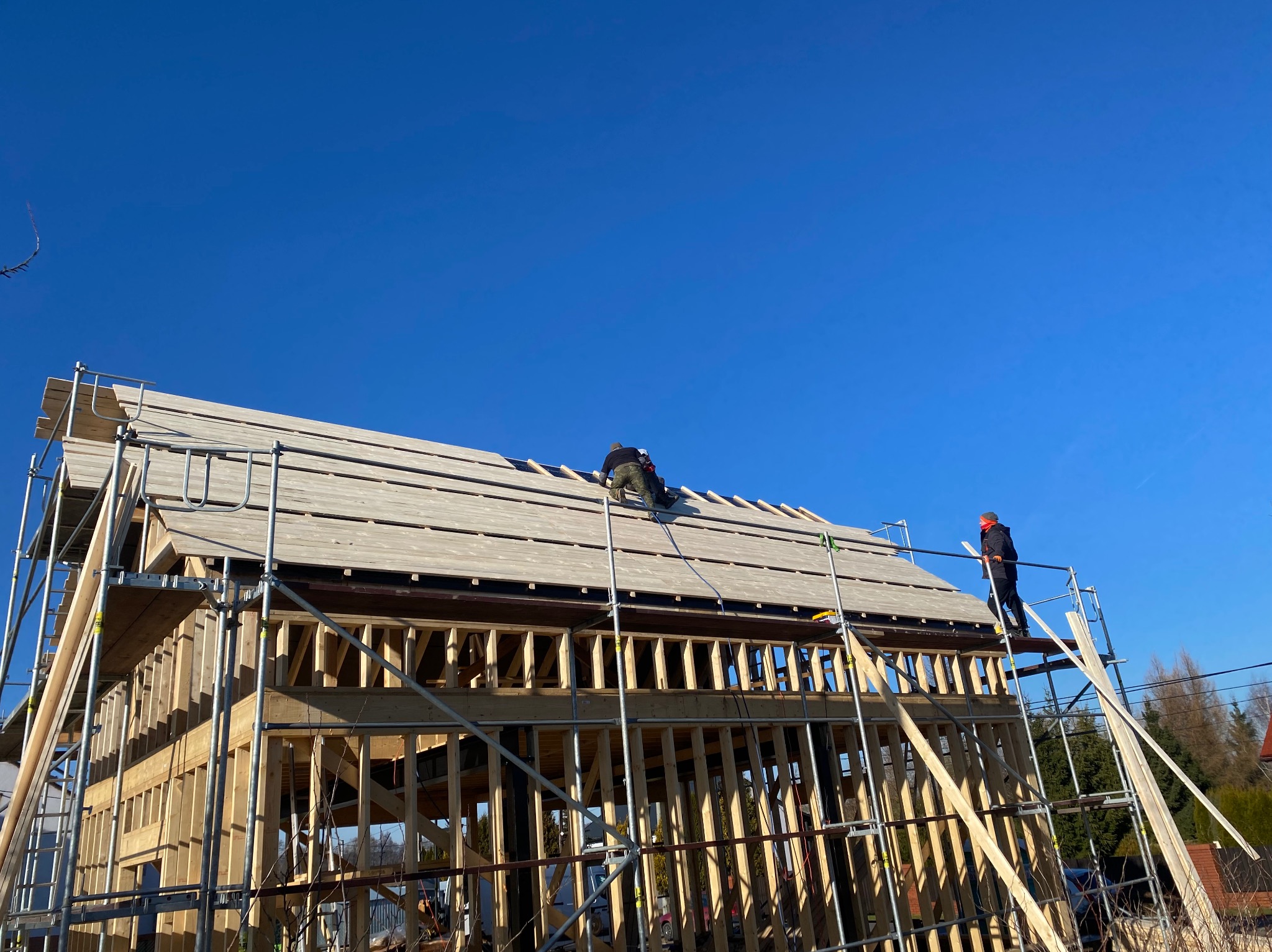 Montaż drewnianej konstrukcji domu szkieletowego z deskowaniem dachu w słoneczny dzień; widoczne rusztowania i dwóch pracowników.