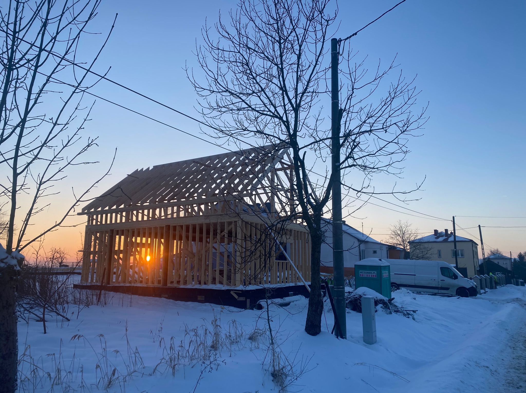 Wczesna faza budowy domu szkieletowego w zimowej scenerii; widoczna drewniana konstrukcja ścian i dachu, z zachodzącym słońcem prześwitującym przez otwory, zasypane śniegiem otoczenie i gołe drzewa...