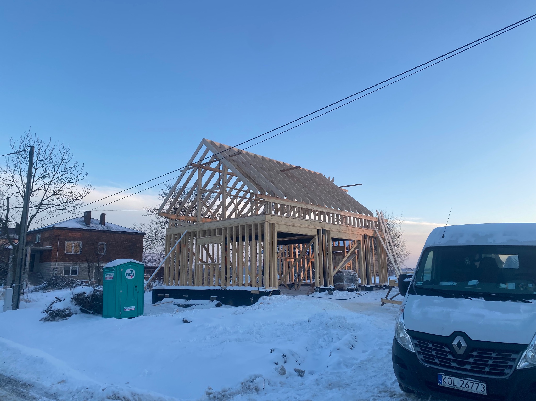 Wznoszenie szkieletu drewnianego domu w zimowej scenerii, widoczny przenośny WC i samochód dostawczy na pierwszym planie.