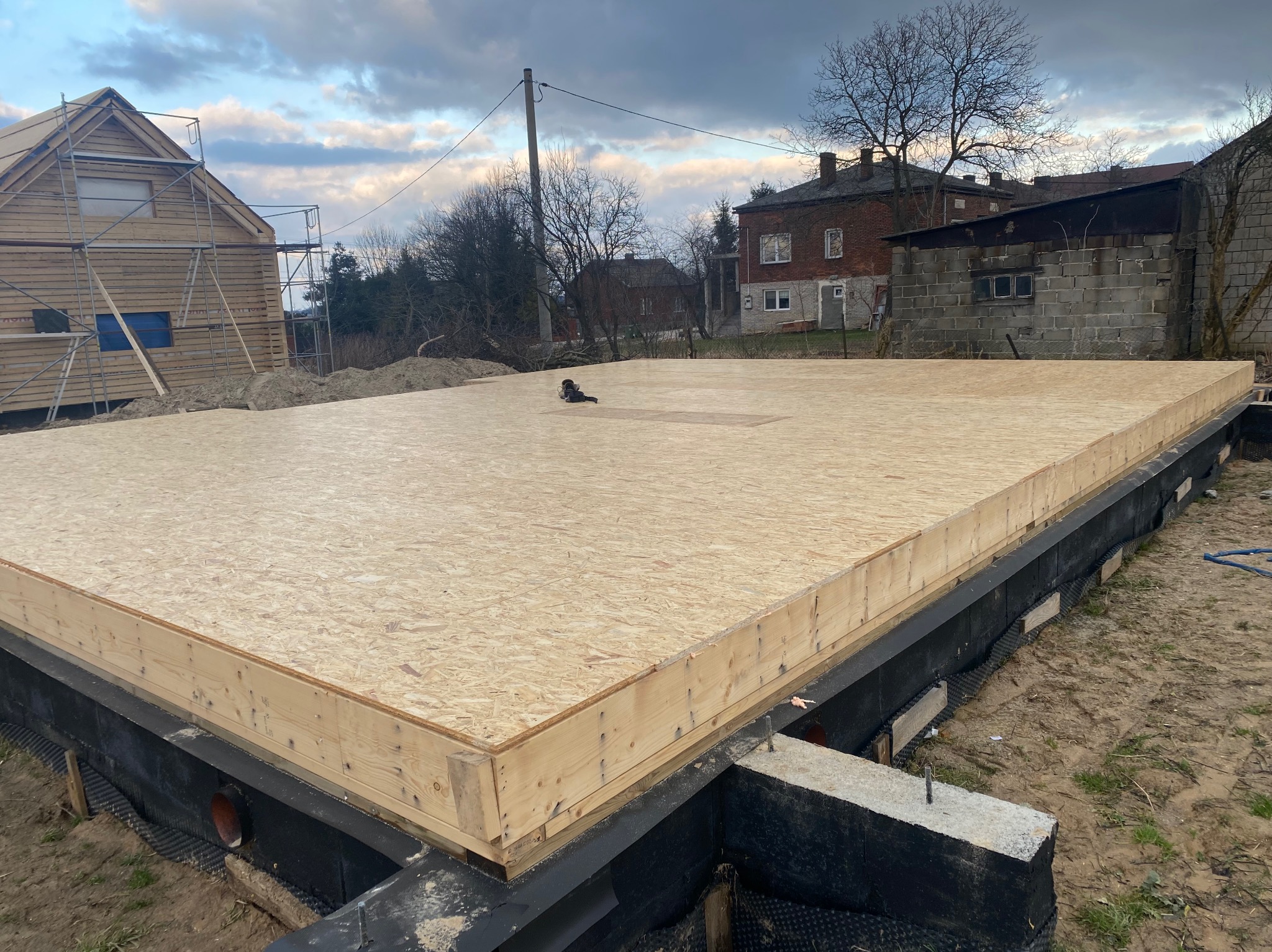 Płyta fundamentowa z drewna i OSB na izolowanej podmurówce pod budowę domu, widok z góry, w tle dom w trakcie budowy i zabudowania gospodarcze.