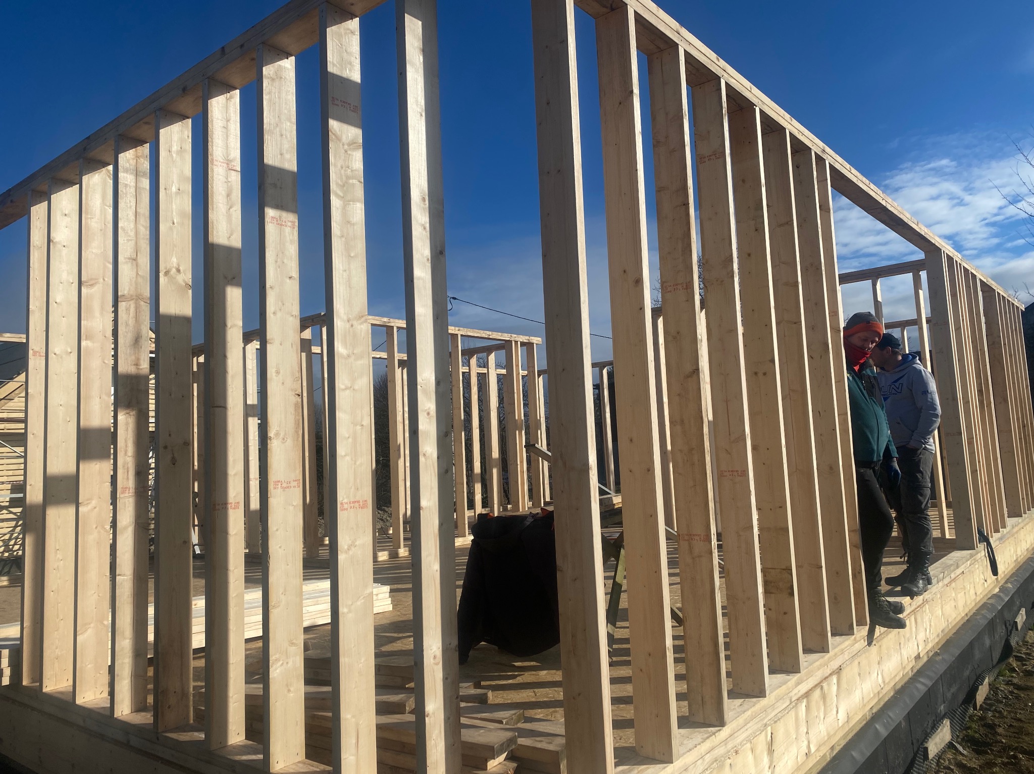 Wznoszenie szkieletu drewnianego domu z widocznymi elementami konstrukcyjnymi i dwoma pracownikami w tle, błękitne niebo w tle.