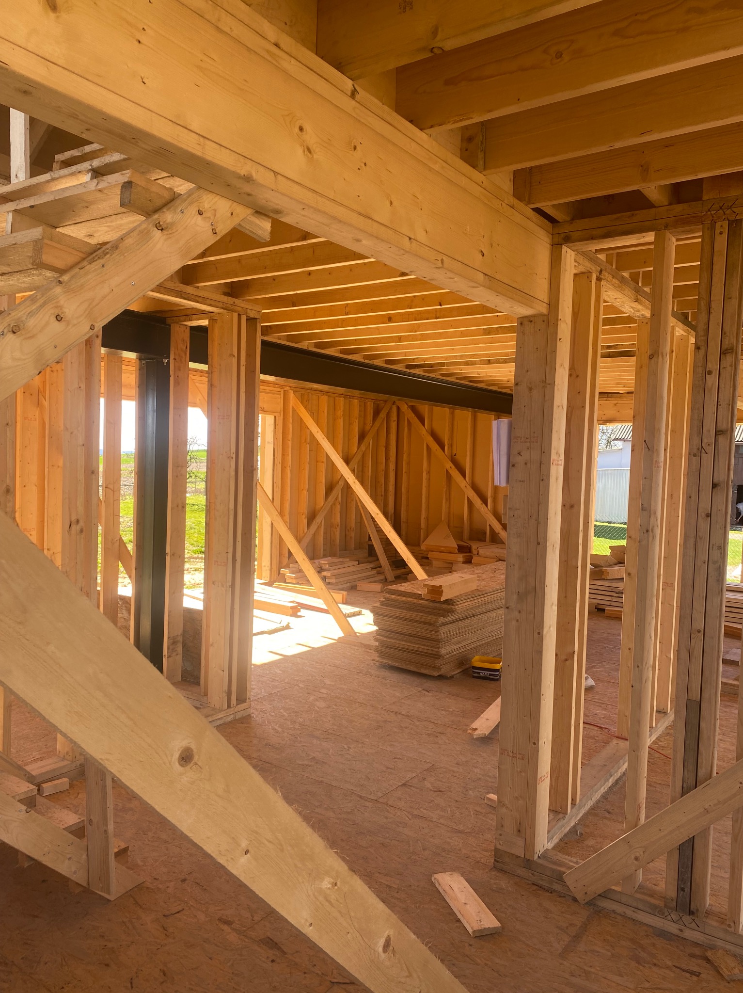 Wnętrze domu w trakcie budowy szkieletowej, widoczne drewniane elementy konstrukcyjne, podłoga z płyt OSB, w tle widok na zewnątrz przez otwór okienny.