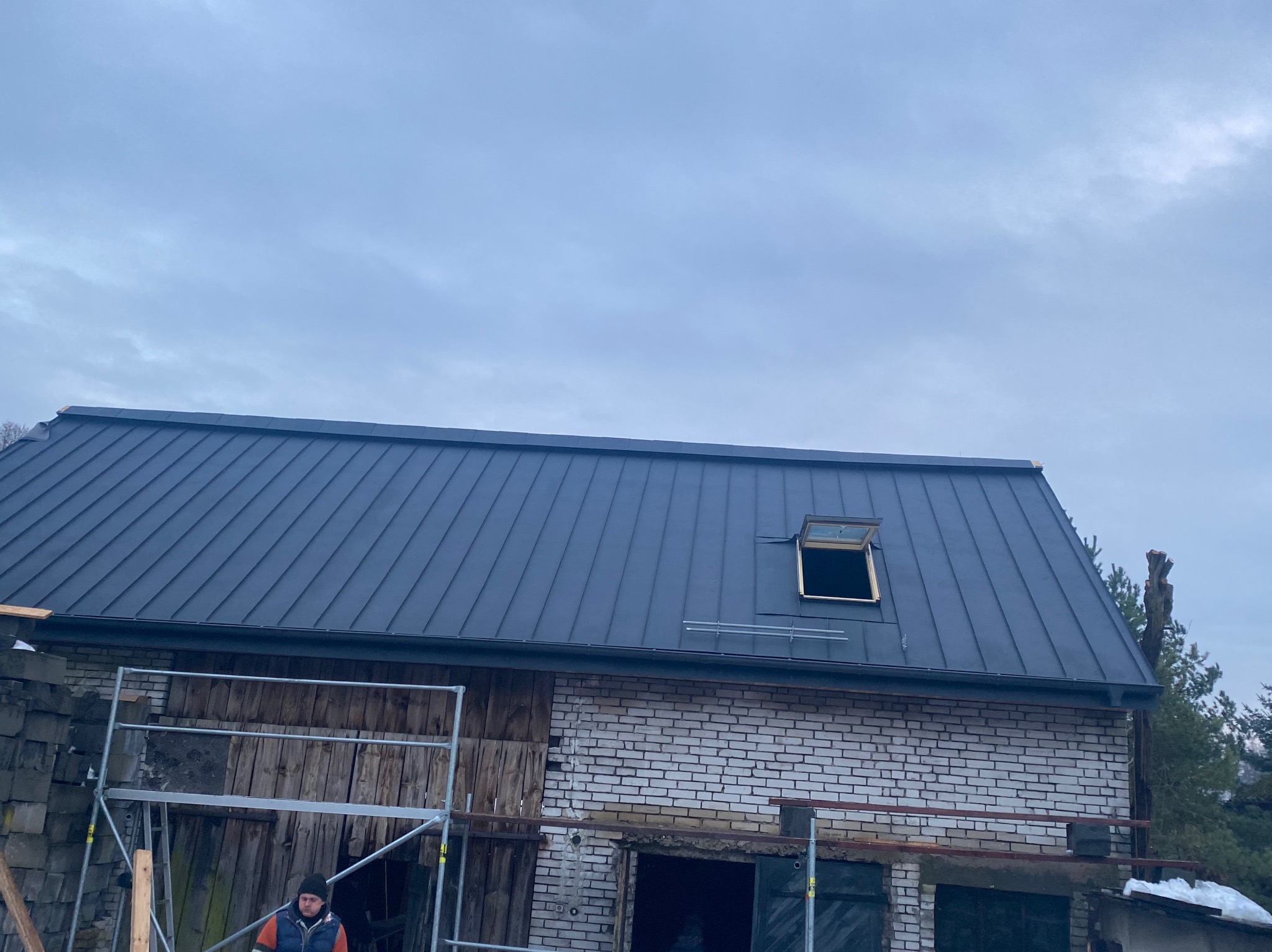 Budynek z nowym, ciemnym dachem z blachy na rąbek stojący, otwartym oknem dachowym, fragmentem rusztowania i pracownikiem w dole, w tle pochmurne niebo.
