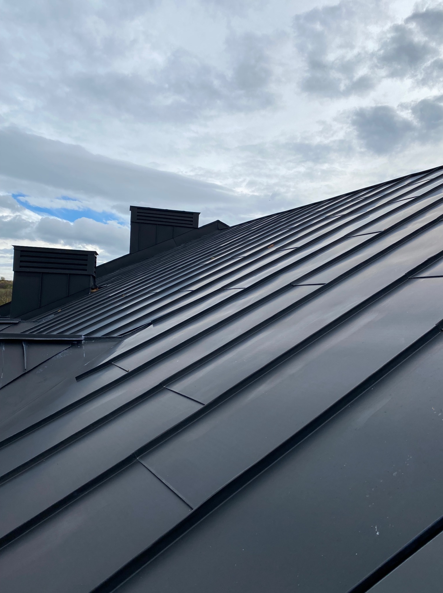 Nowoczesny, ciemny dach z blachy na tle pochmurnego nieba, widoczne kominy wentylacyjne.