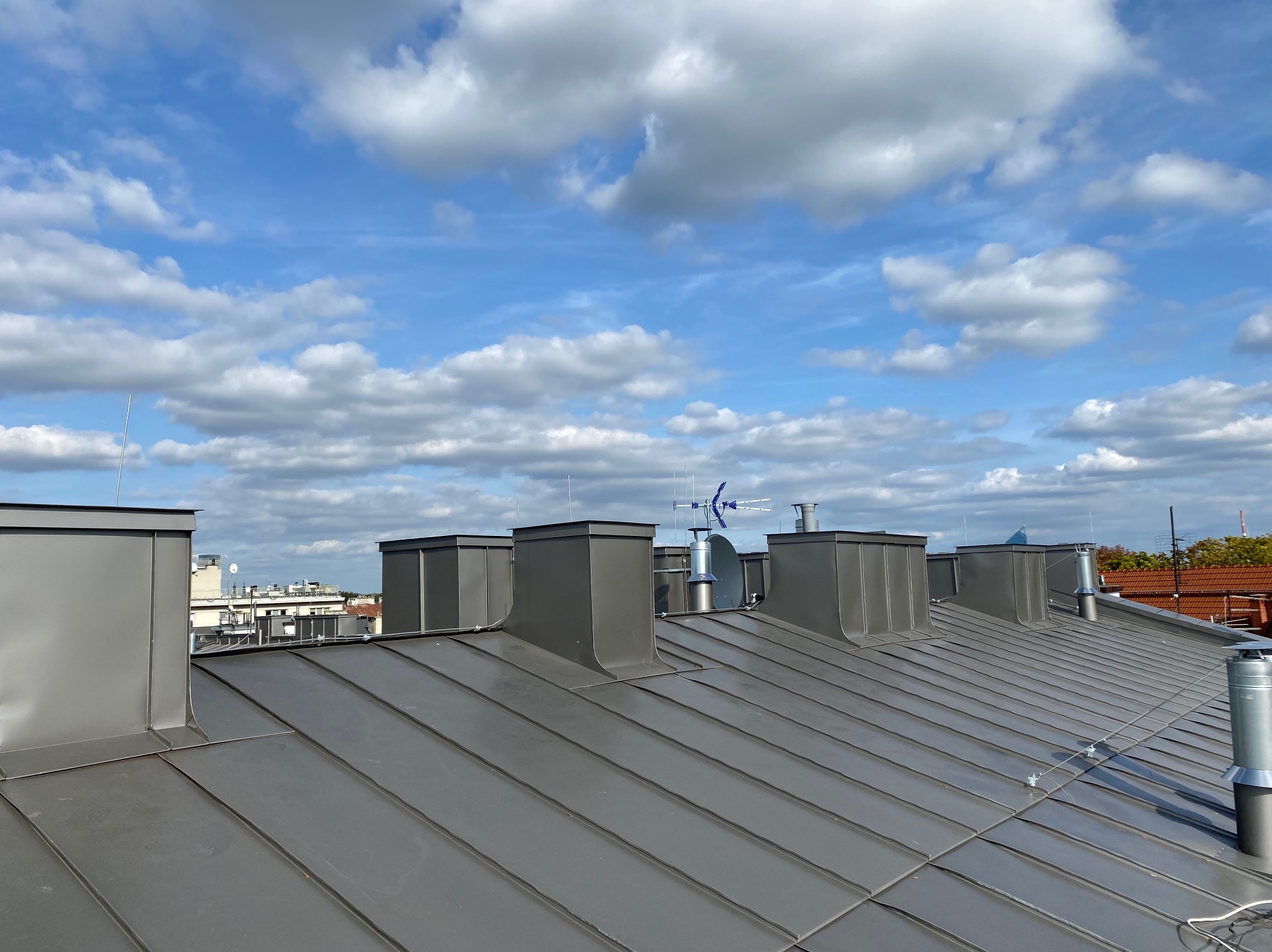 Szary metalowy dach z kominami i anteną satelitarną na tle błękitnego nieba z obłokami.