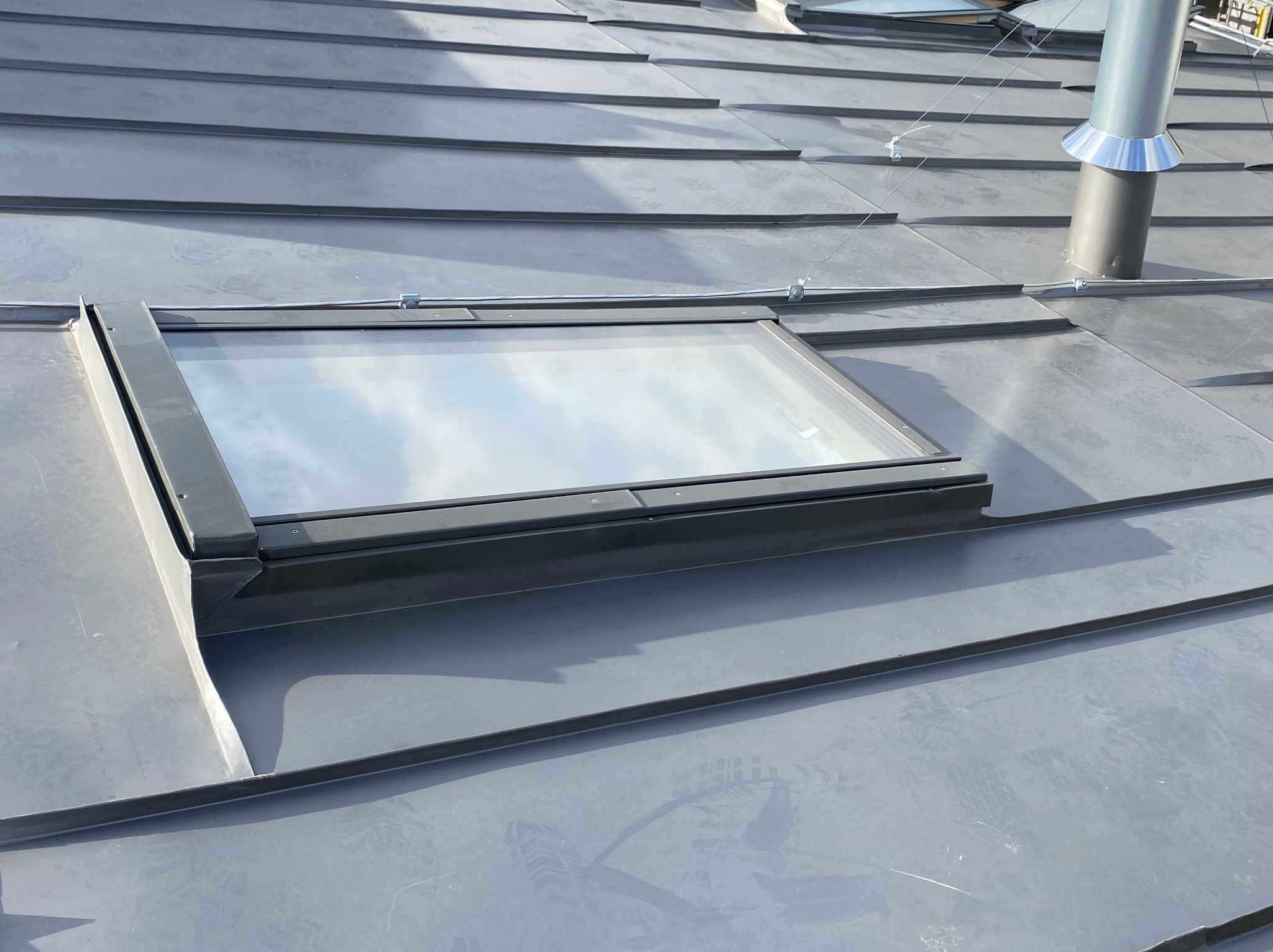 Szary metalowy dach z oknem dachowym i kominem wentylacyjnym, widoczne mocowania i odbicie nieba w szybie.