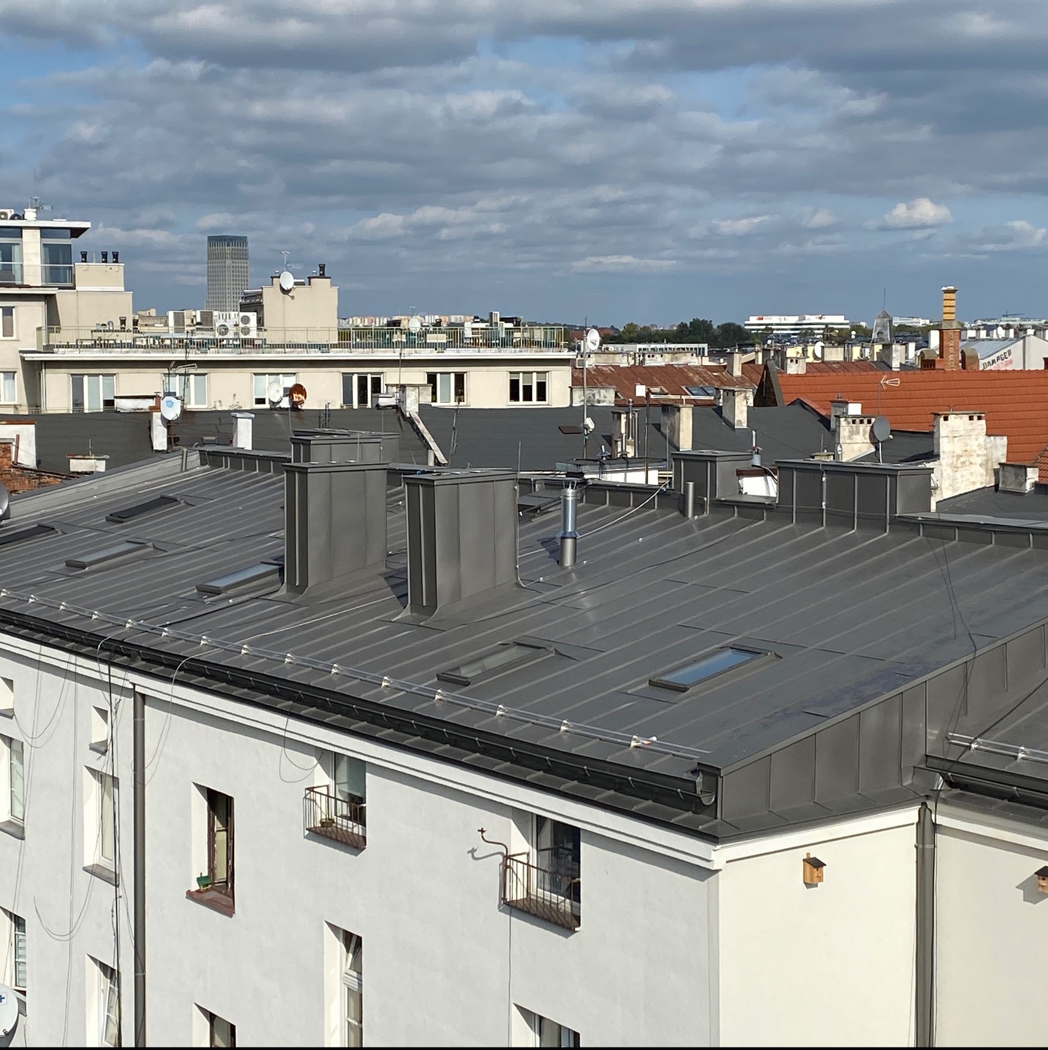 Szary metalowy dach z lukarnami i kominami wentylacyjnymi na tle miejskiego krajobrazu z budynkami i pochmurnym niebem.