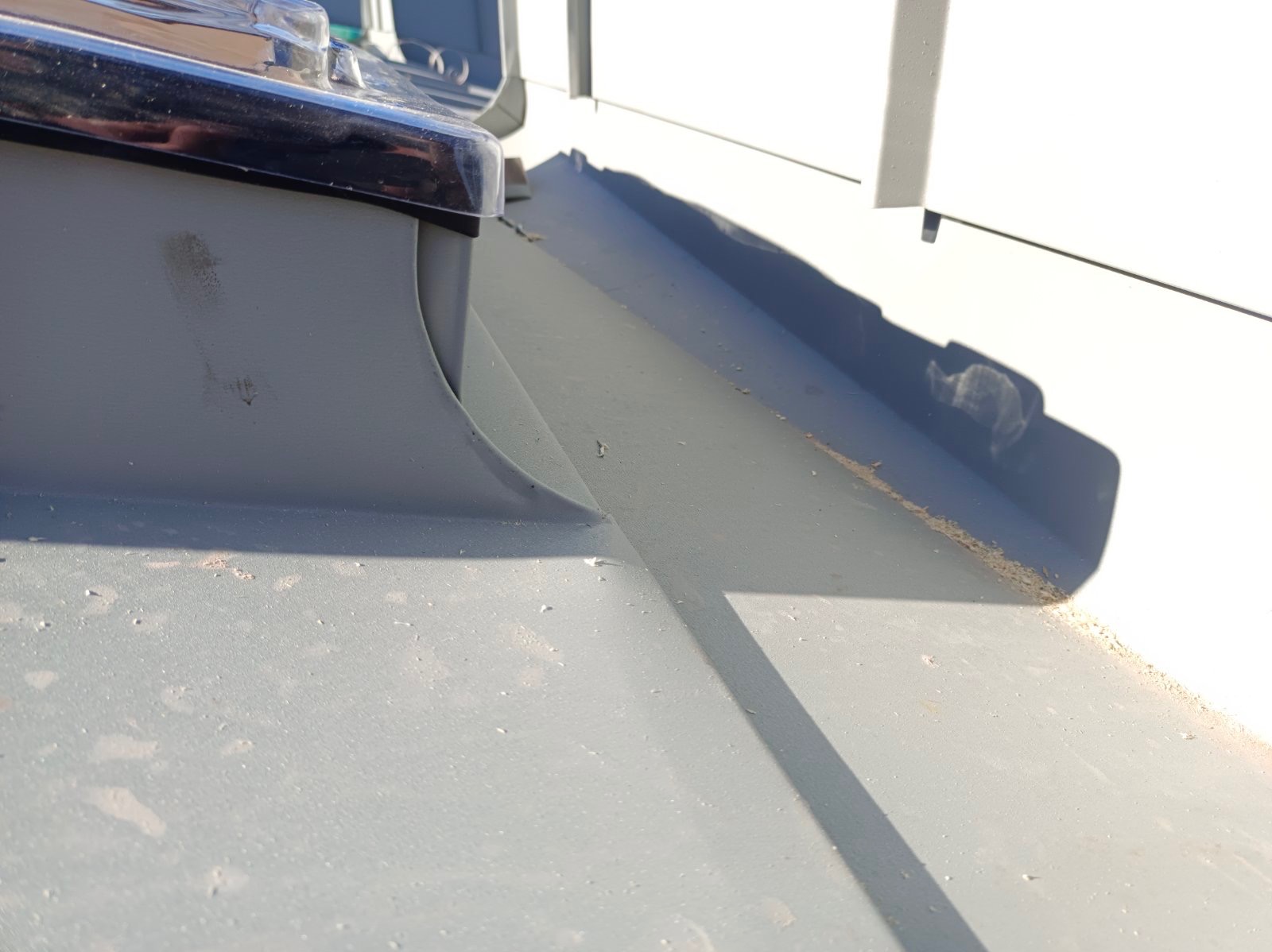 Szary metalowy element dachu z widocznym łączeniem blach i śladami drobnych zabrudzeń, oświetlony jasnym światłem słonecznym.