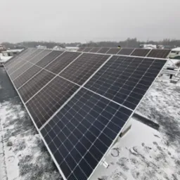 Panele słoneczne na dachu pokrytym śniegiem, widok z góry na instalację na tle zabudowań mieszkalnych w pochmurny dzień.