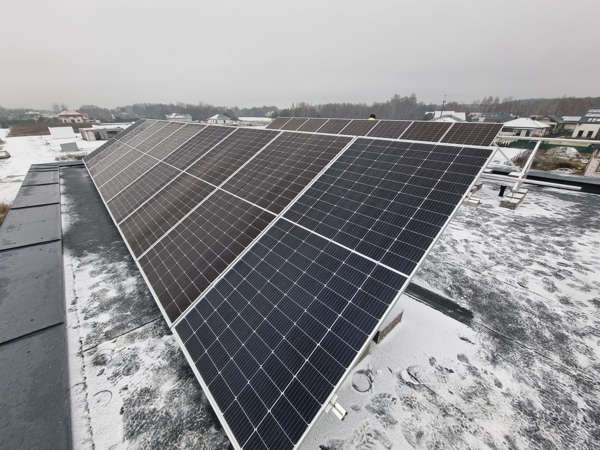 Panele słoneczne na dachu pokrytym śniegiem, widok z góry na instalację na tle zabudowań mieszkalnych w pochmurny dzień.