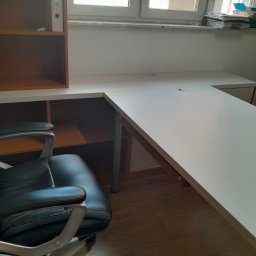 Meble Andrii Pavlenko - Białe biurko narożne z blatem z otworami na kable, przylegające do brązowego regału z półkami, obok okna i krzesła biurowego.