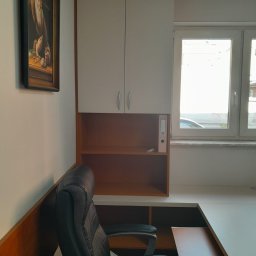 Meble Andrii Pavlenko - Białe szafki wiszące nad biurkiem z brązowymi półkami, czarne krzesło biurowe i obraz na ścianie w tle okna.