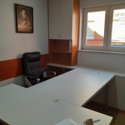 Meble Andrii Pavlenko - Biuro z białym, narożnym biurkiem, czarnym fotelem, szafkami wiszącymi i obrazem na ścianie