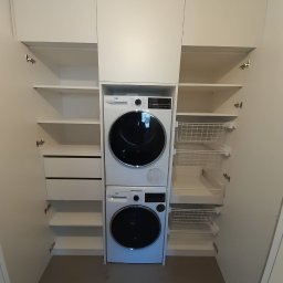 Meble Andrii Pavlenko - Biała szafa na wymiar z zabudowaną pralką i suszarką, półki i wysuwane kosze na pranie po bokach.