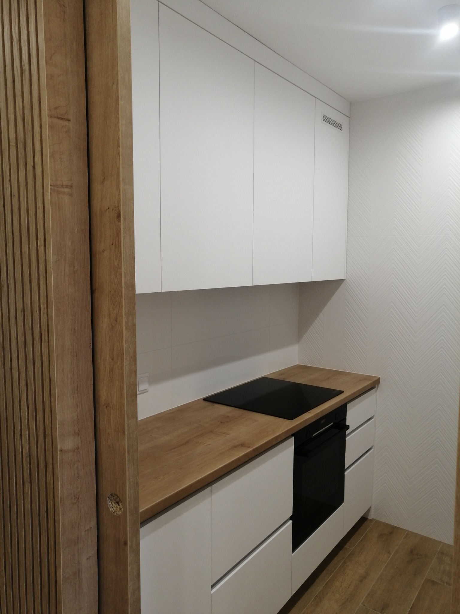 Nowoczesna kuchnia: białe szafki z drewnianym blatem, płyta indukcyjna i piekarnik, drewniane panele dekoracyjne na ścianie.