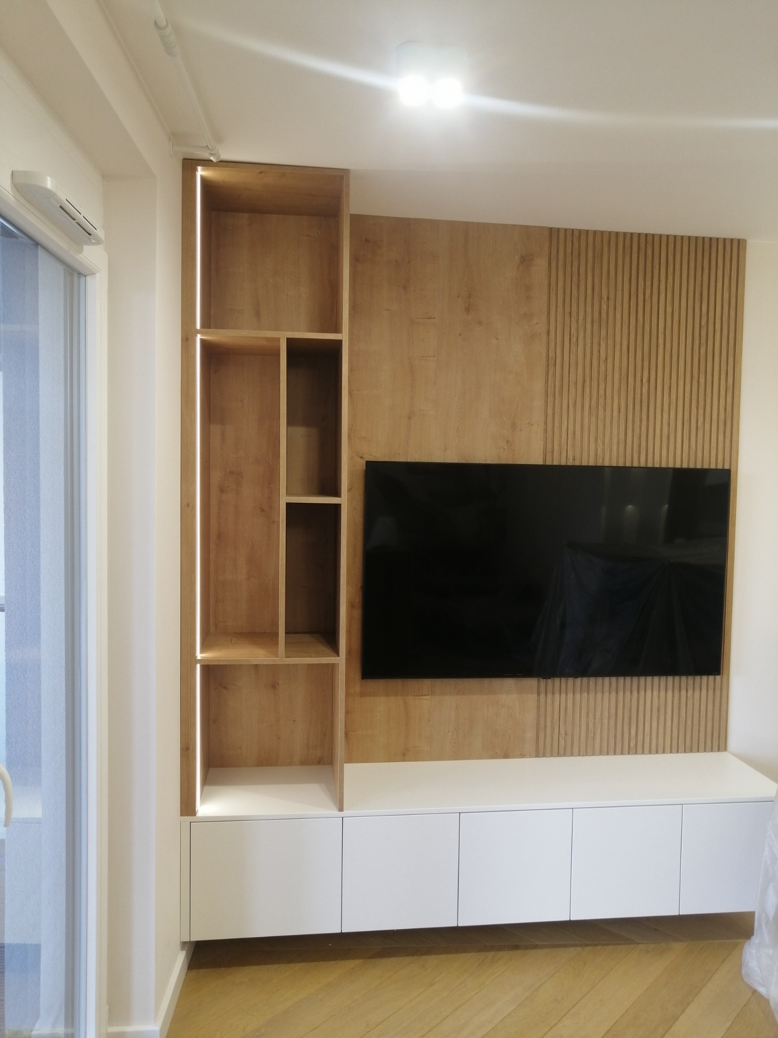 Nowoczesna zabudowa ściany z telewizorem: drewniane panele pionowe, regał z podświetleniem LED i biała szafka RTV z frontami bez uchwytów.