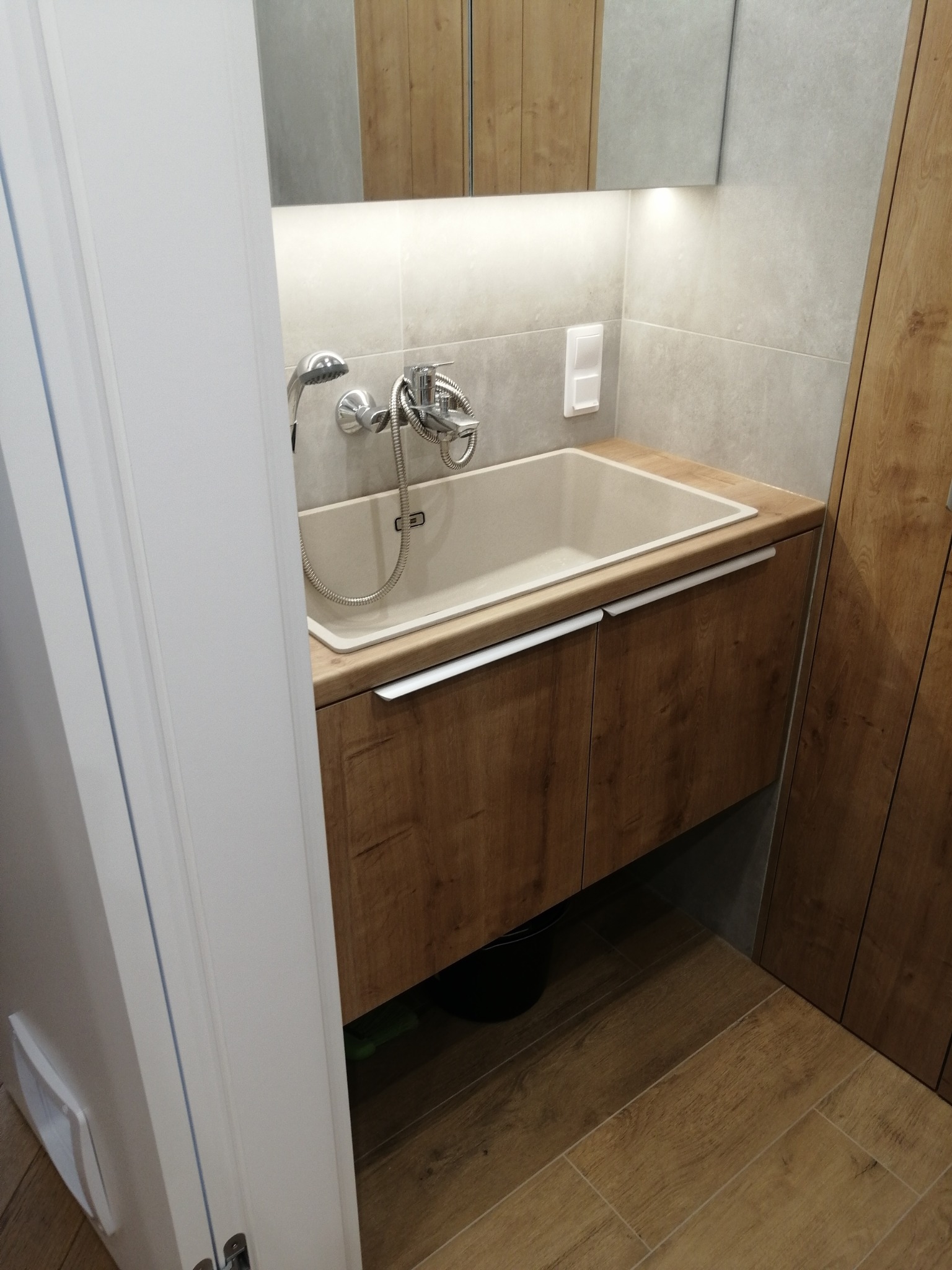 Nowoczesna łazienka: umywalka nablatowa z drewnianą szafką, szara ściana z podświetlanym lustrem, drewniana podłoga, widoczny fragment drzwi.