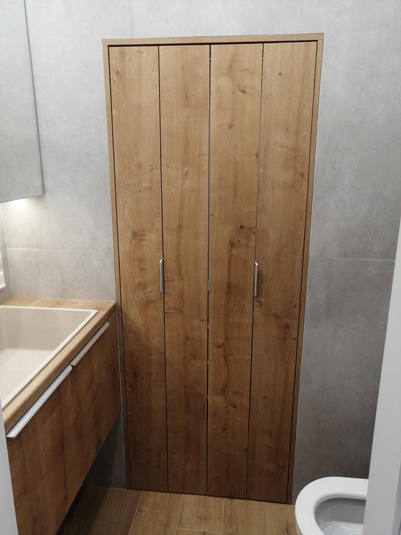 Czterodrzwiowa szafa wnękowa z drewnopodobnymi frontami w łazience, z widoczną umywalką i fragmentem toalety.