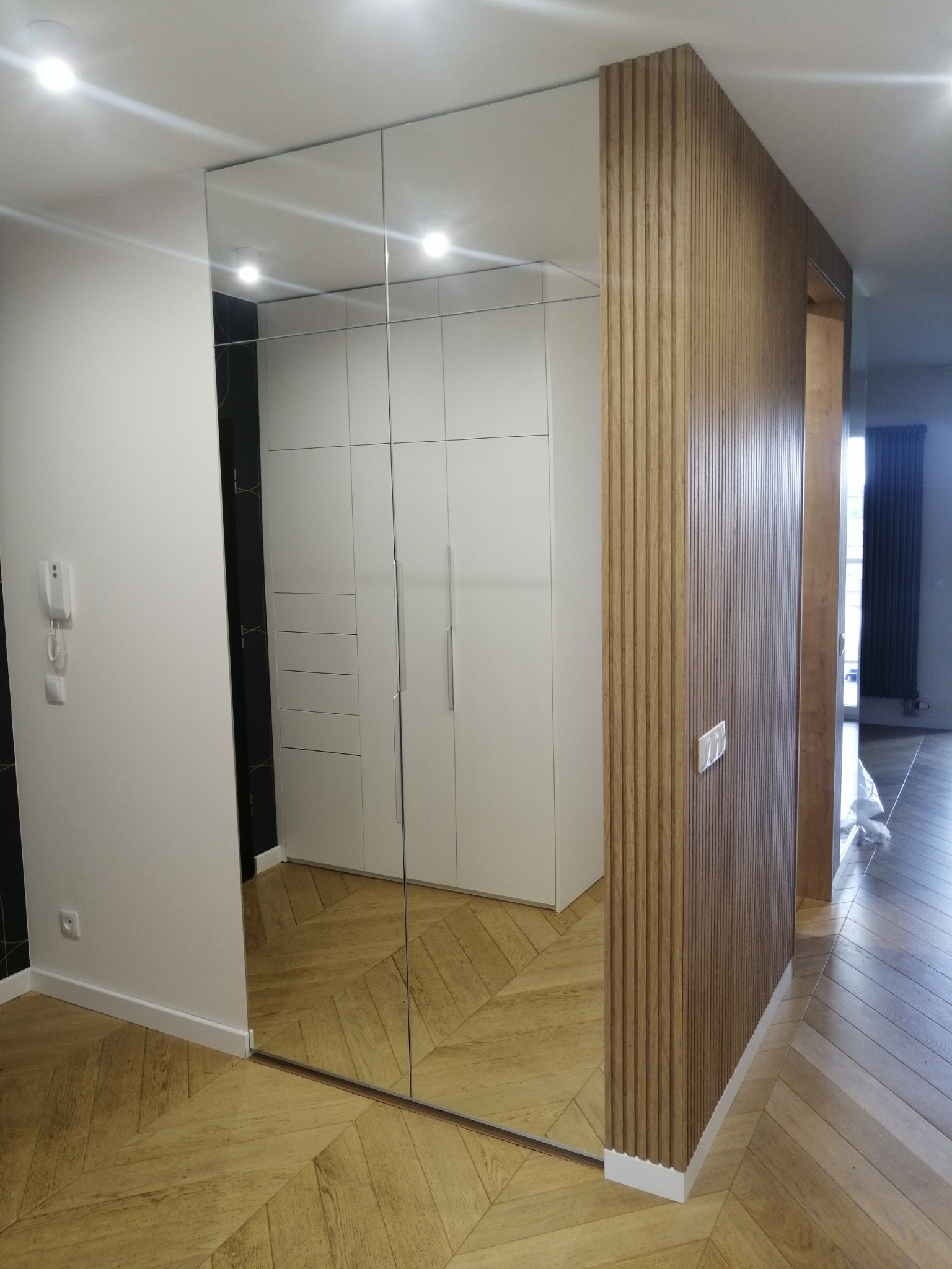 Nowoczesna szafa wnękowa z lustrzanymi drzwiami przesuwnymi, odbijająca wnętrze z drewnianą podłogą w jodełkę i ścianą z drewnianych paneli.