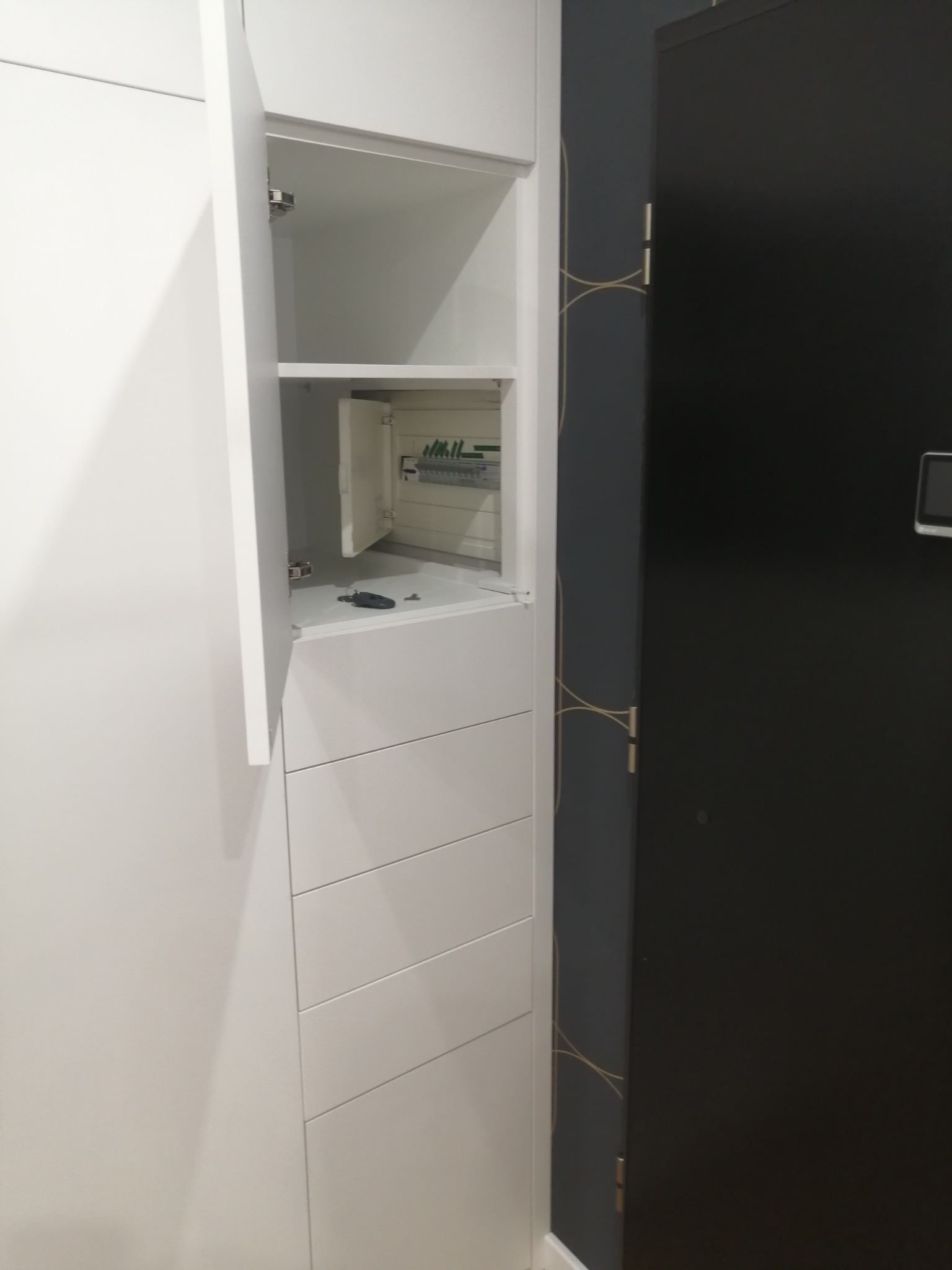 Biała szafa wnękowa z otwartą szafką, w której widoczna jest skrzynka z bezpiecznikami i klucze.