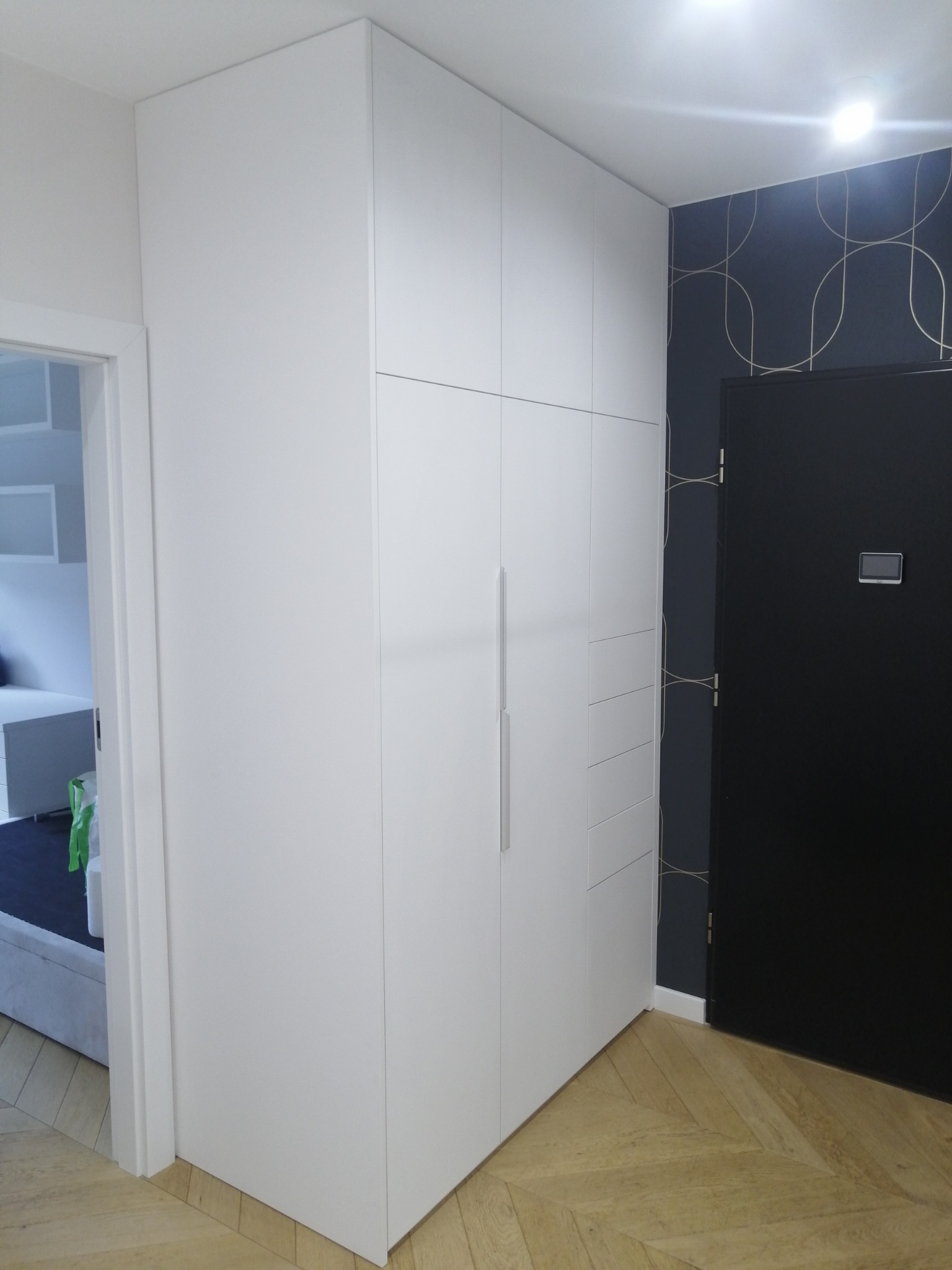 Biała szafa wnękowa z długim, pionowym uchwytem, w tle czarne drzwi i ściana z geometrycznym wzorem, widok z perspektywy podłogi z parkietem w jodełkę.