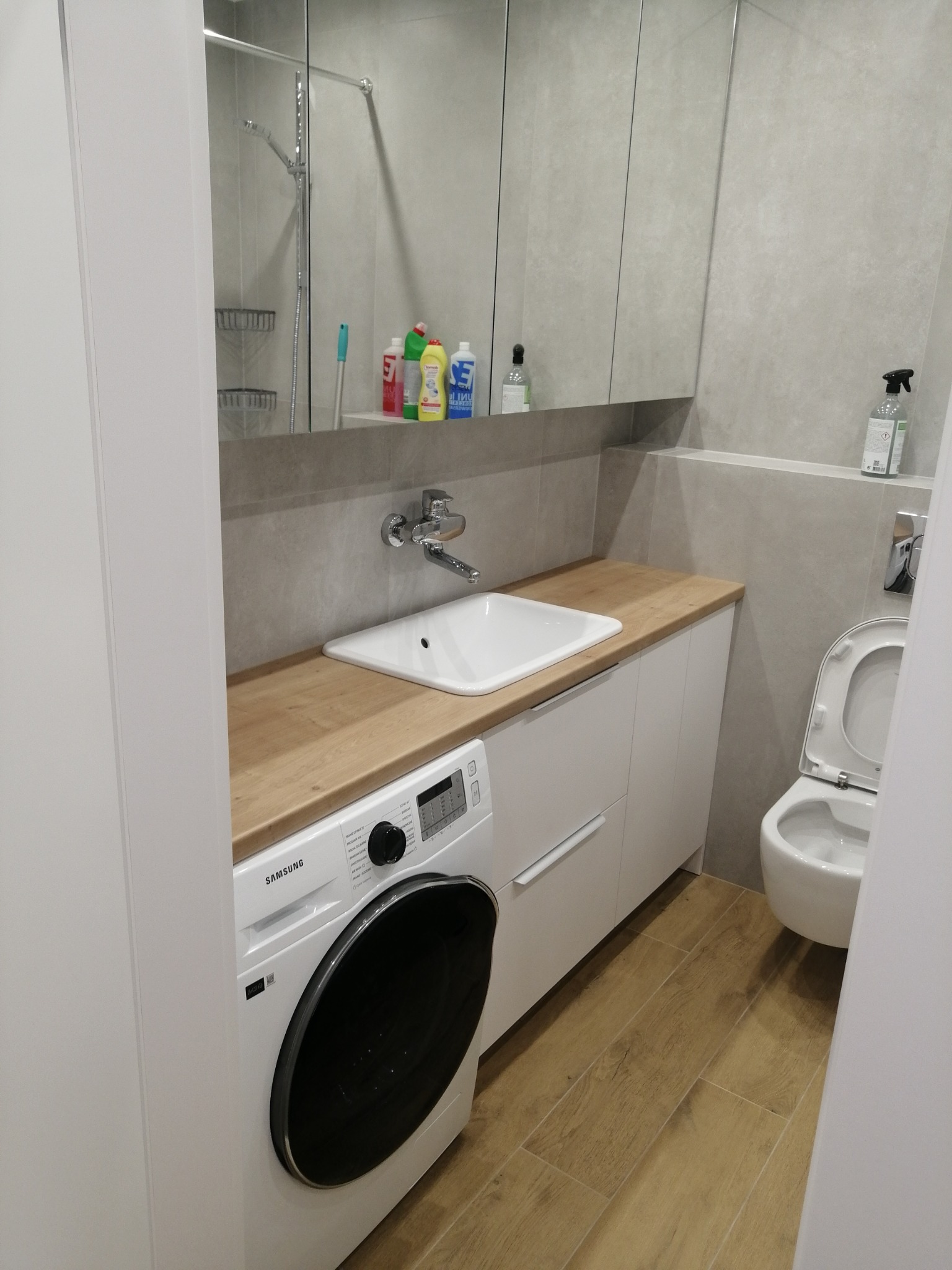 Nowoczesna łazienka z drewnianym blatem, białą umywalką, pralką Samsung i szafką z lustrem, minimalistyczny design.
