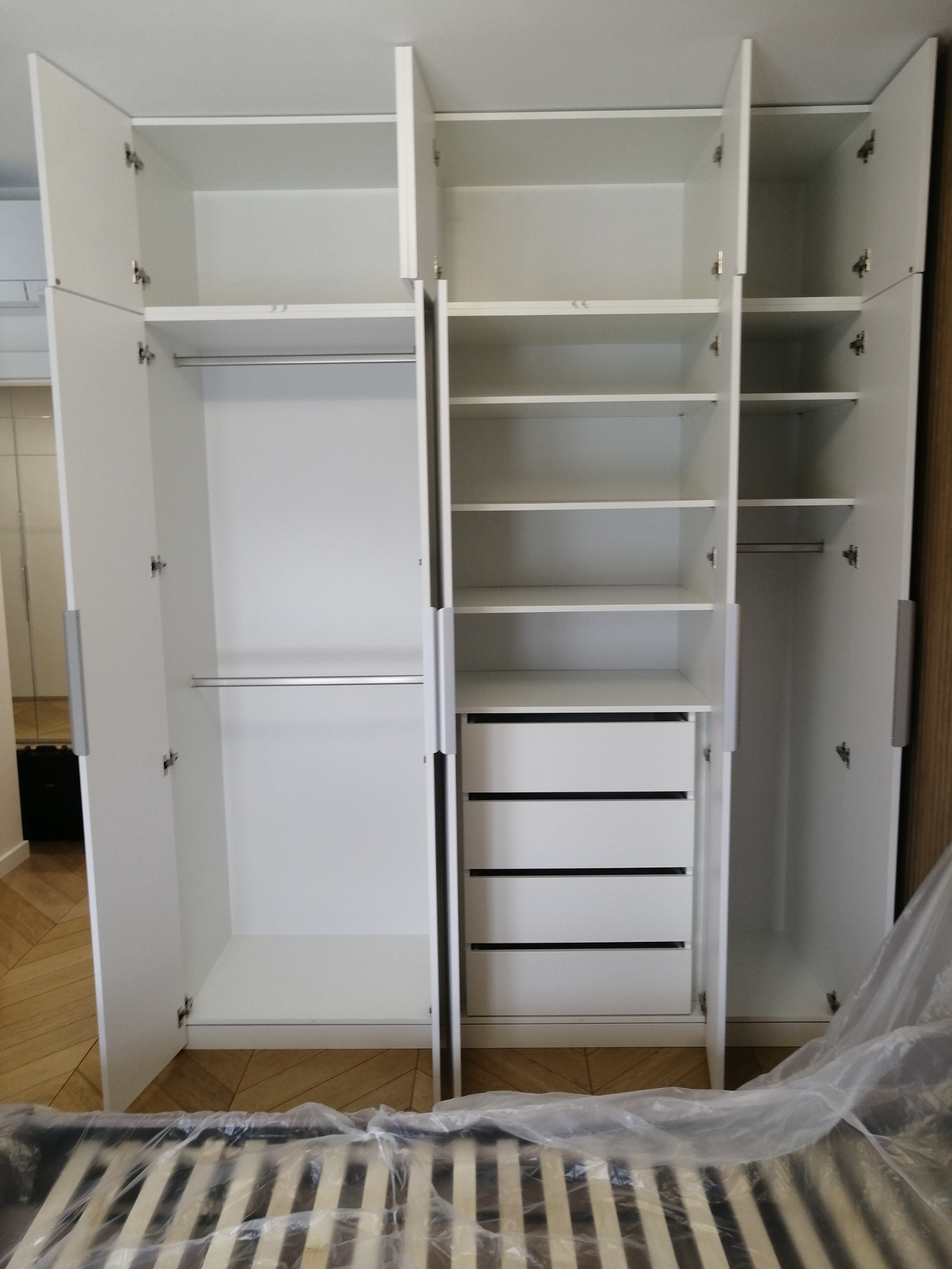 Wnętrze białej, trzydrzwiowej szafy z półkami, drążkami i szufladami, widoczna częściowo rama łóżka zabezpieczona folią.
