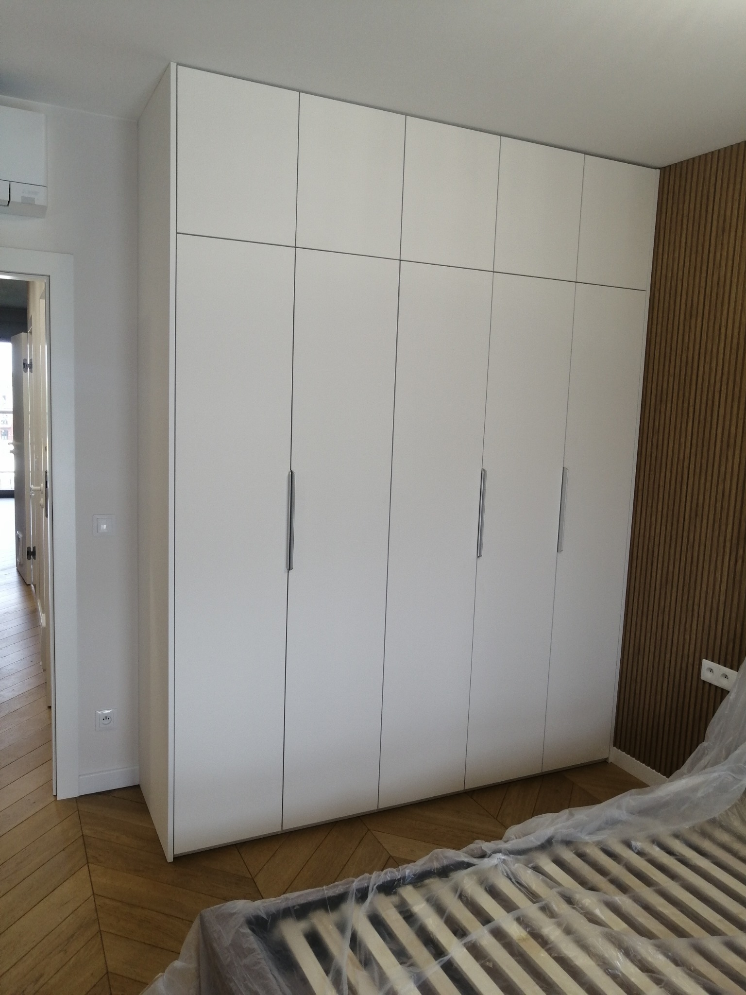 Biała szafa wnękowa z minimalistycznymi uchwytami, dopasowana do drewnianej boazerii na ścianie. Widoczny fragment łóżka w folii ochronnej i parkiet w jodełkę.