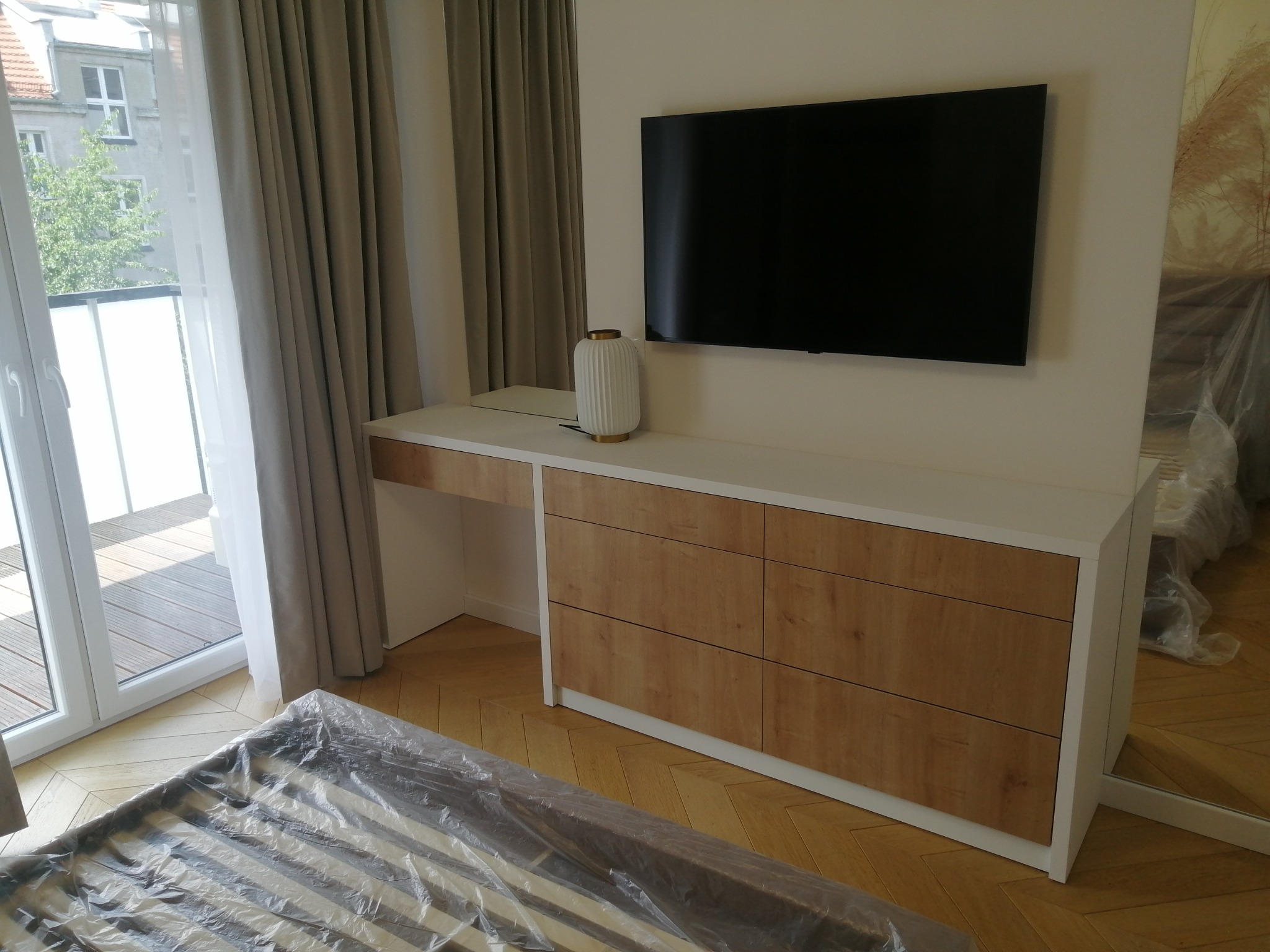 Szafka na wymiar z drewnianymi frontami szuflad i białym blatem, wkomponowana pod telewizor, z lampą dekoracyjną, w tle balkon i parkiet w jodełkę, łóżko w folii ochronnej.