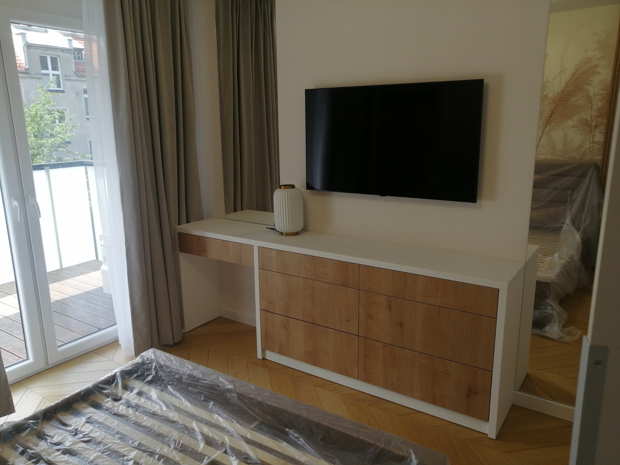Nowoczesna szafka RTV z biurkiem w kolorze białym i drewnopodobnym, z telewizorem na ścianie, widok na balkon i częściowo zakryte łóżko folią ochronną.