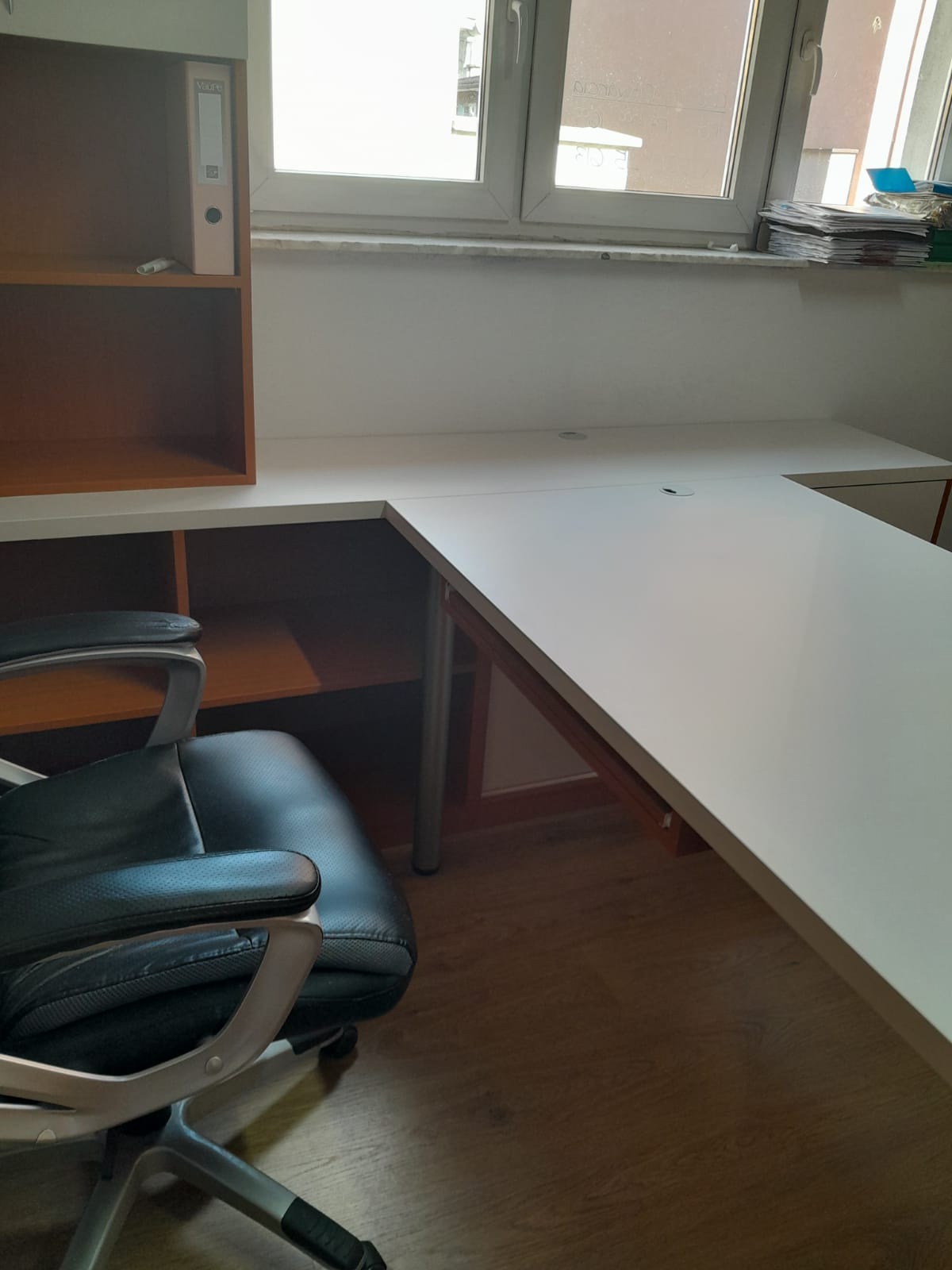 Białe biurko narożne z blatem z otworami na kable, przylegające do brązowego regału z półkami, obok okna i krzesła biurowego.