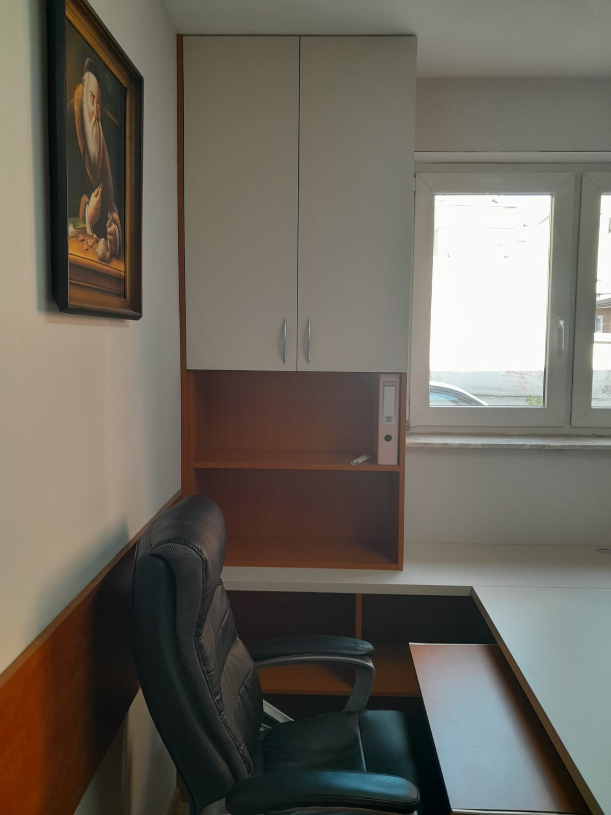 Białe szafki wiszące nad biurkiem z brązowymi półkami, czarne krzesło biurowe i obraz na ścianie w tle okna.