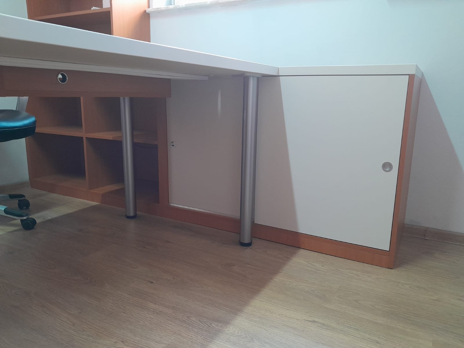 Fragment biurka narożnego z szafką i półkami, wspartego na metalowych nogach, widoczne z perspektywy podłogi.