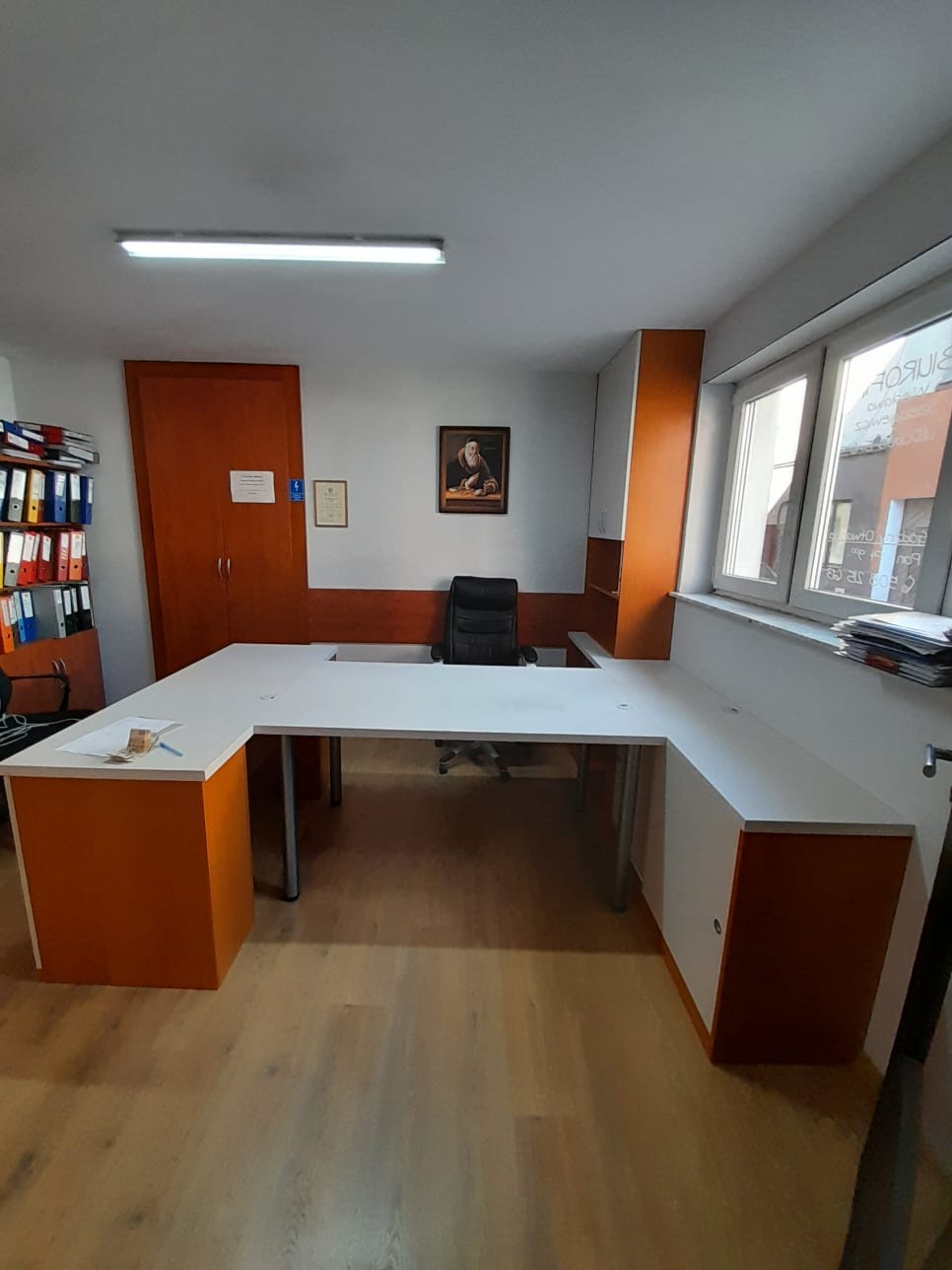 Białe biurko narożne z pomarańczowymi akcentami, w tle regał z segregatorami i portret na ścianie w biurze.