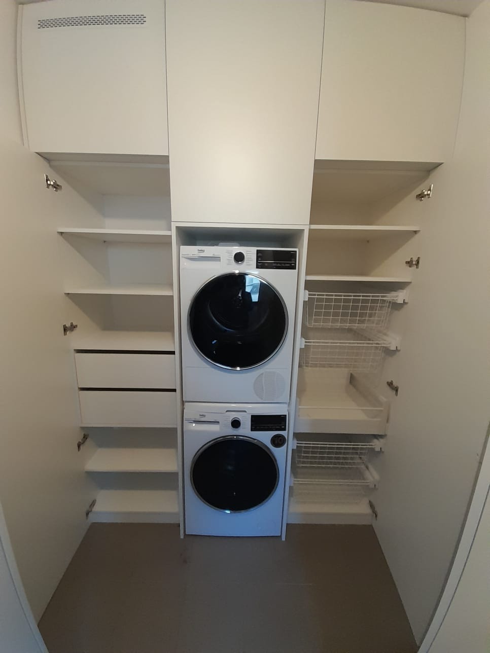 Biała szafa na wymiar z zabudowaną pralką i suszarką, półki i wysuwane kosze na pranie po bokach.