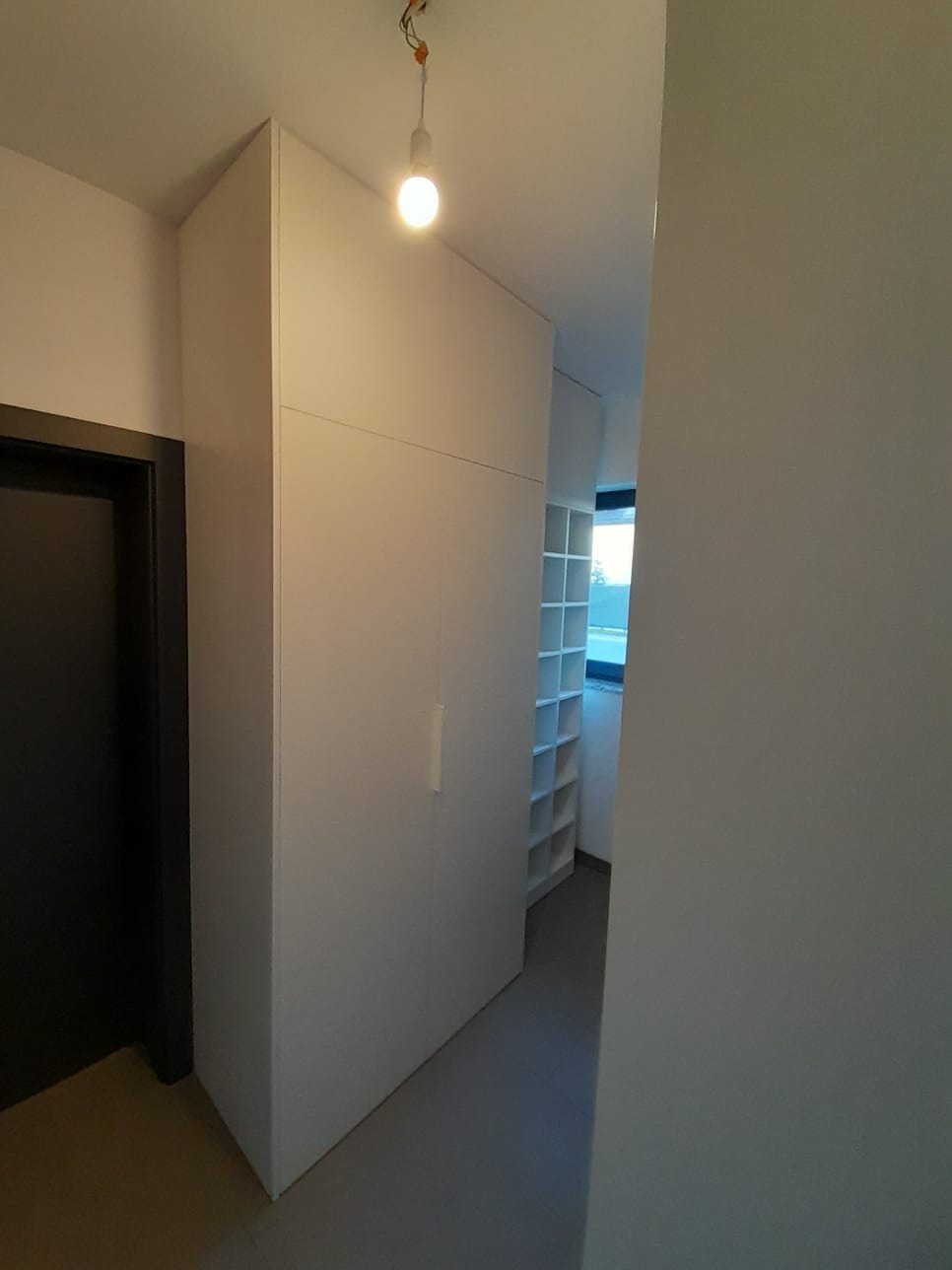 Wysoka, biała szafa wnękowa z długim, pionowym uchwytem, oświetlona żarówką na kablu, widok z korytarza z widocznymi półkami w głębi.