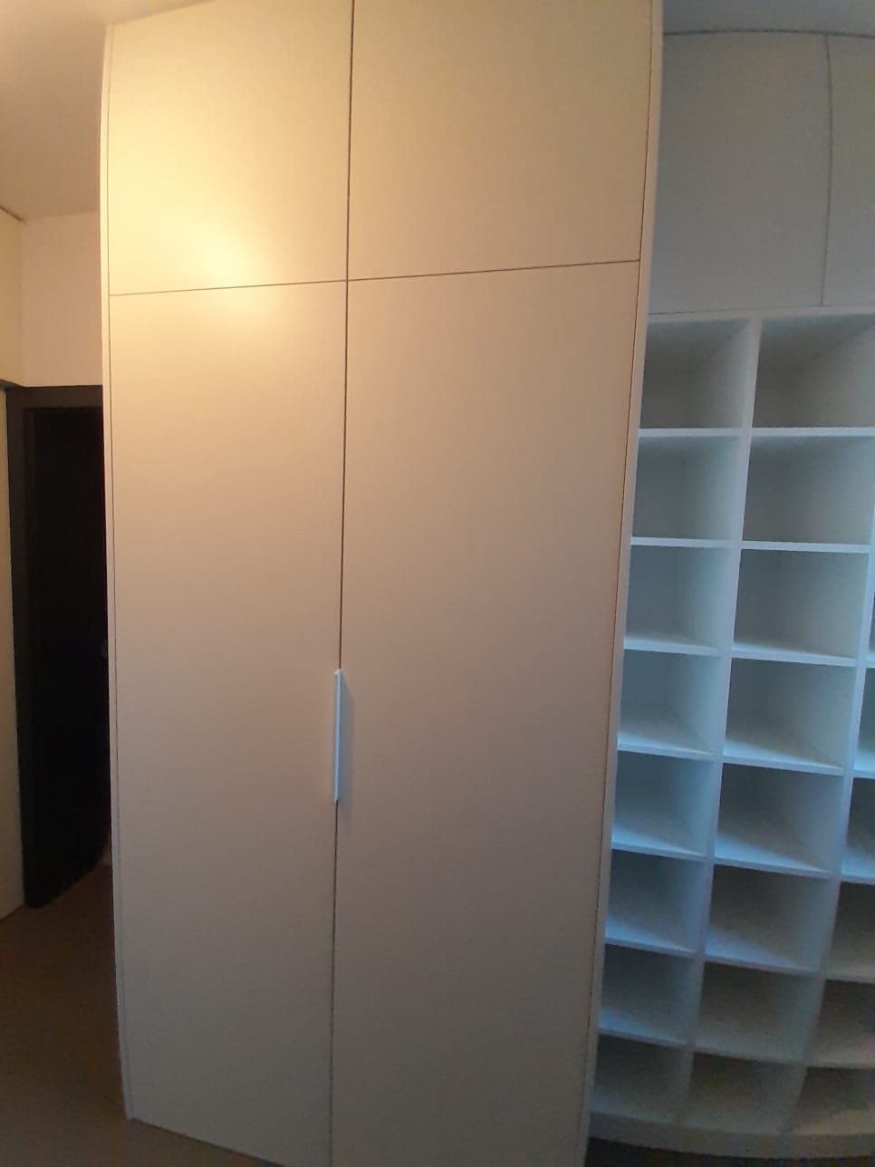 Biała, wysoka szafa z czterema drzwiami i minimalistycznymi uchwytami, obok regał z licznymi kwadratowymi przegródkami, widoczny fragment ciemnego otworu drzwiowego.